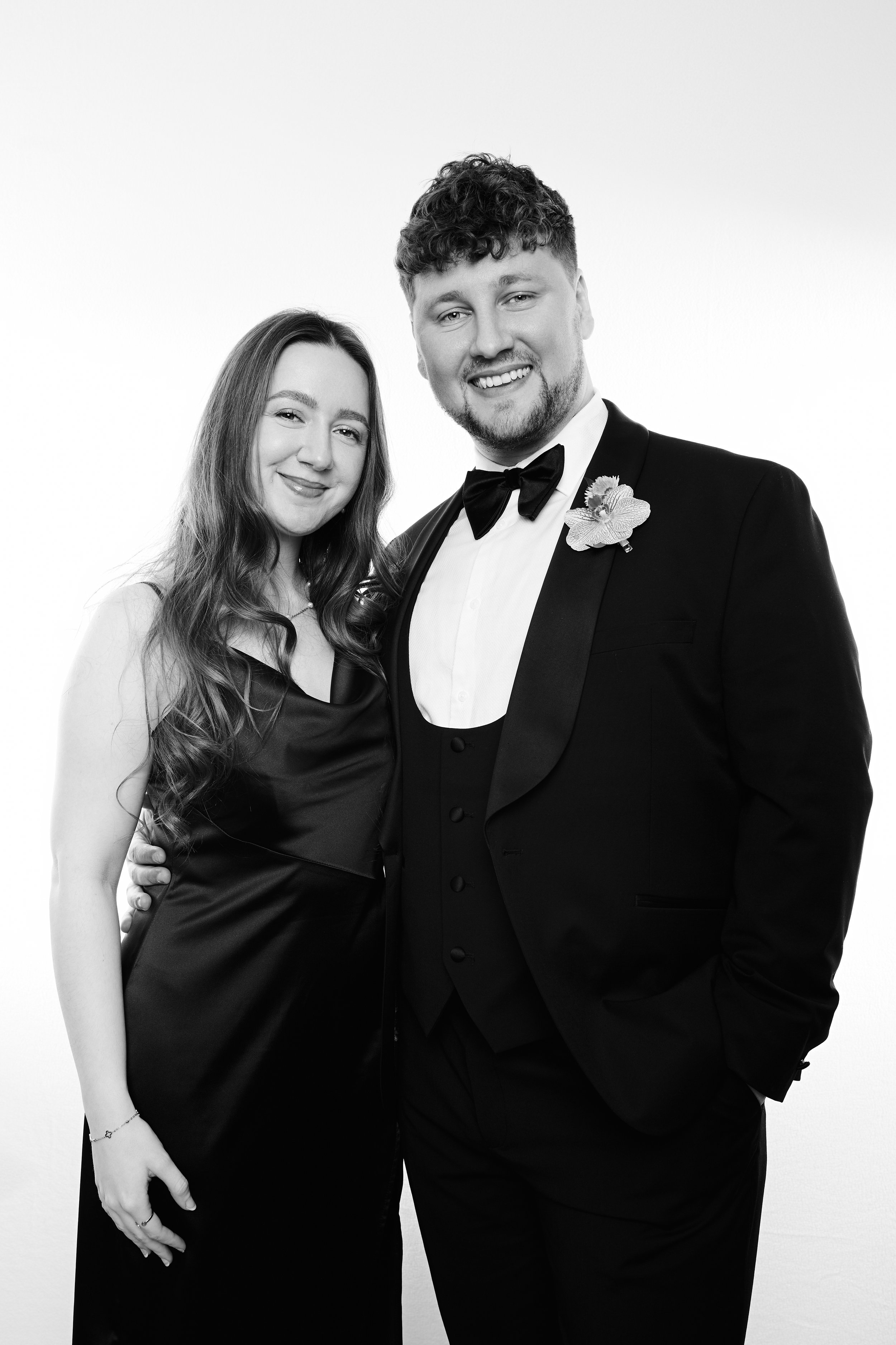 A black-and-white photo of a smiling couple dressed formally in a tuxedo and a satin evening gown, standing close together against a plain light background.