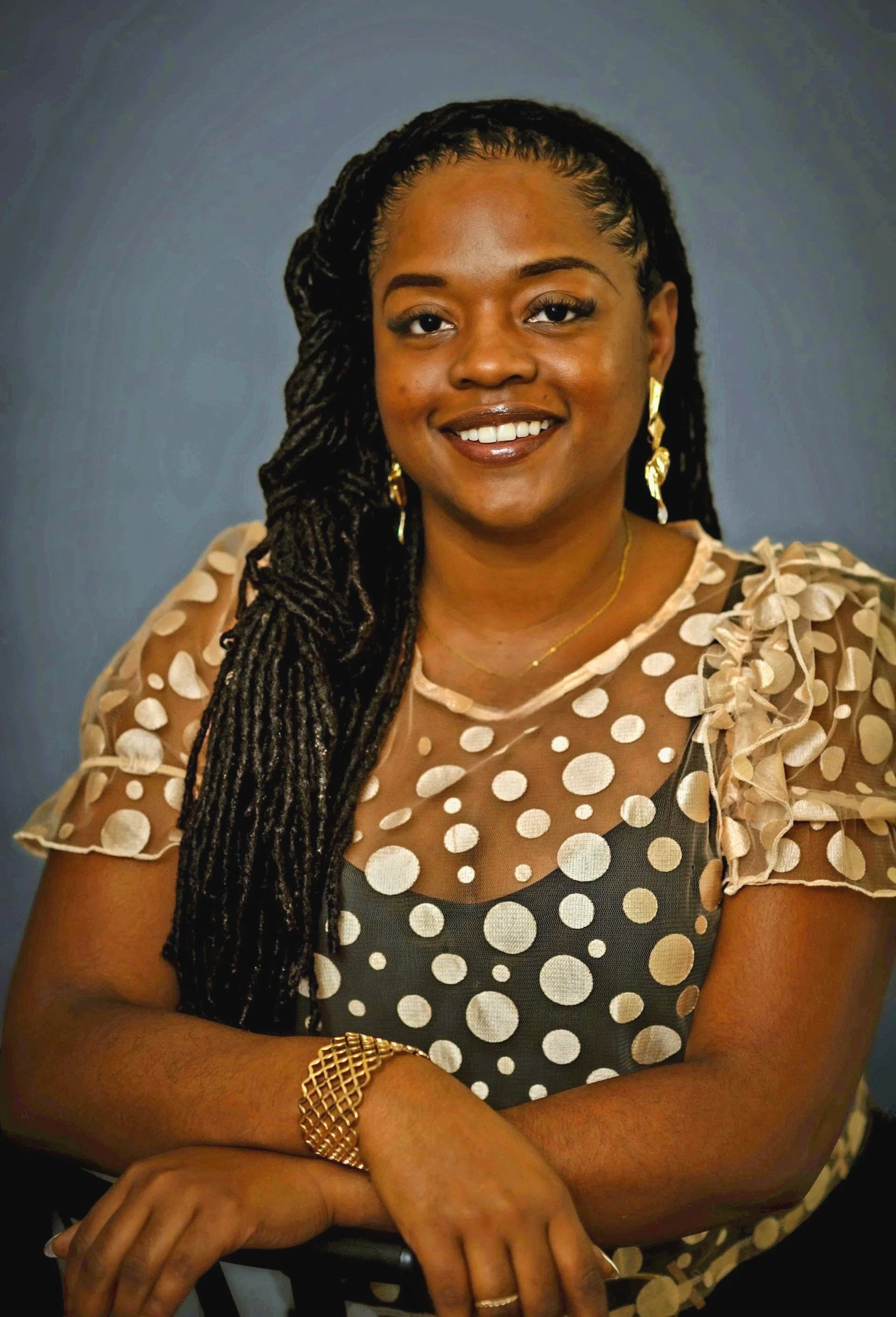 A woman with long twisted hair wearing a peach-colored, polka-dotted sheer top with ruffled sleeves, gold earrings, gold bracelet, and a gold necklace, smiling with arms crossed against a blue background.