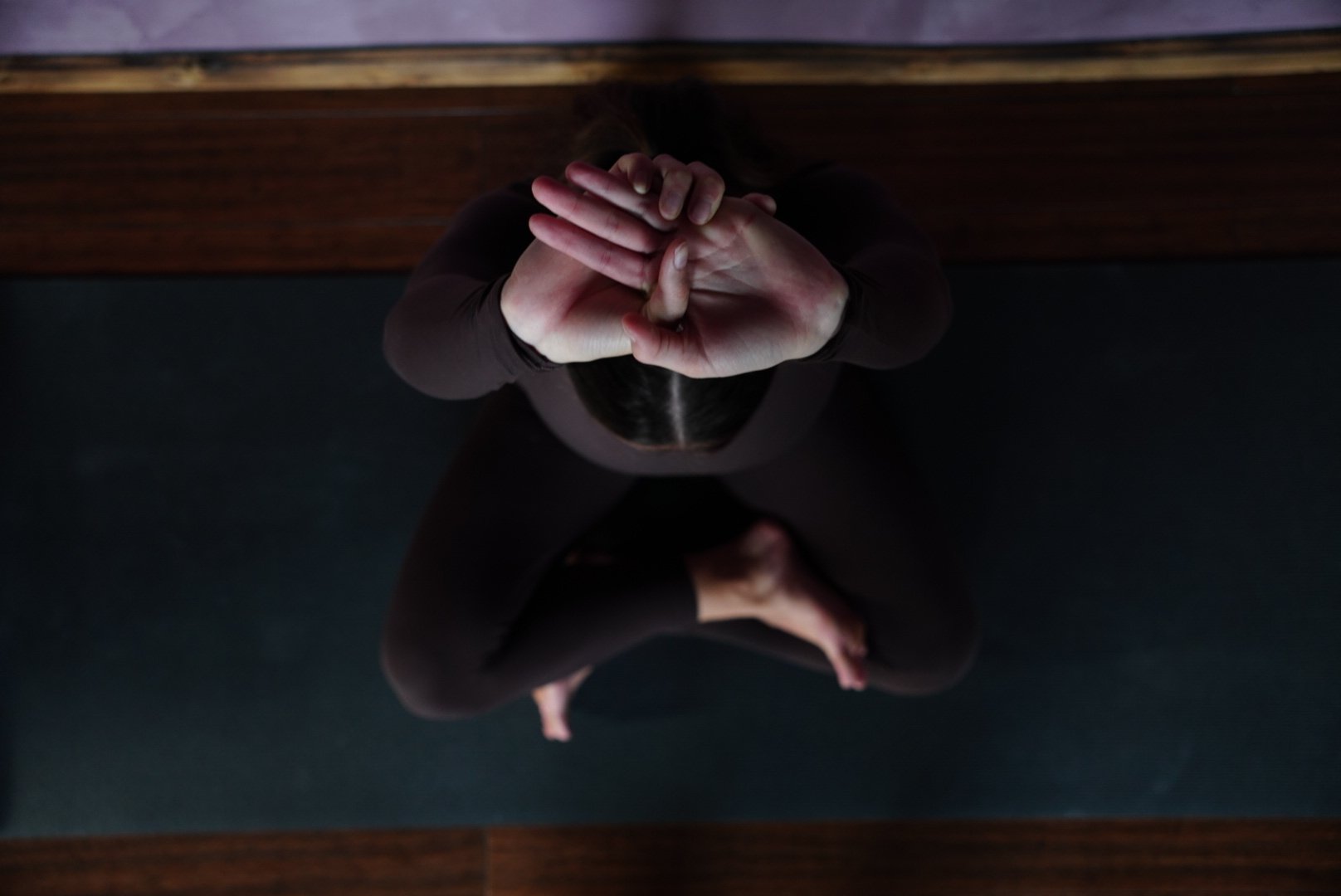 Top-down view of a person sitting cross-legged on the floor with hands pressed together at the forehead in a praying or meditative pose.