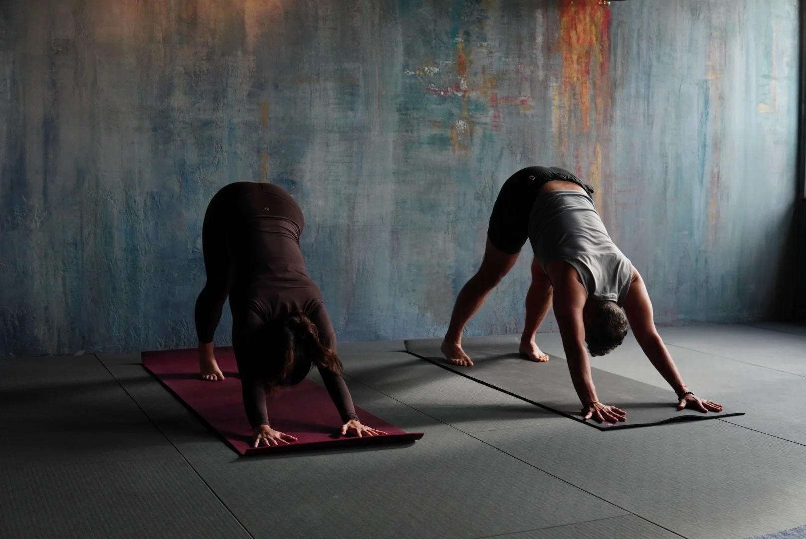 Two people practicing yoga in downward dog pose on mats in a dimly lit room with a colorful abstract wall.