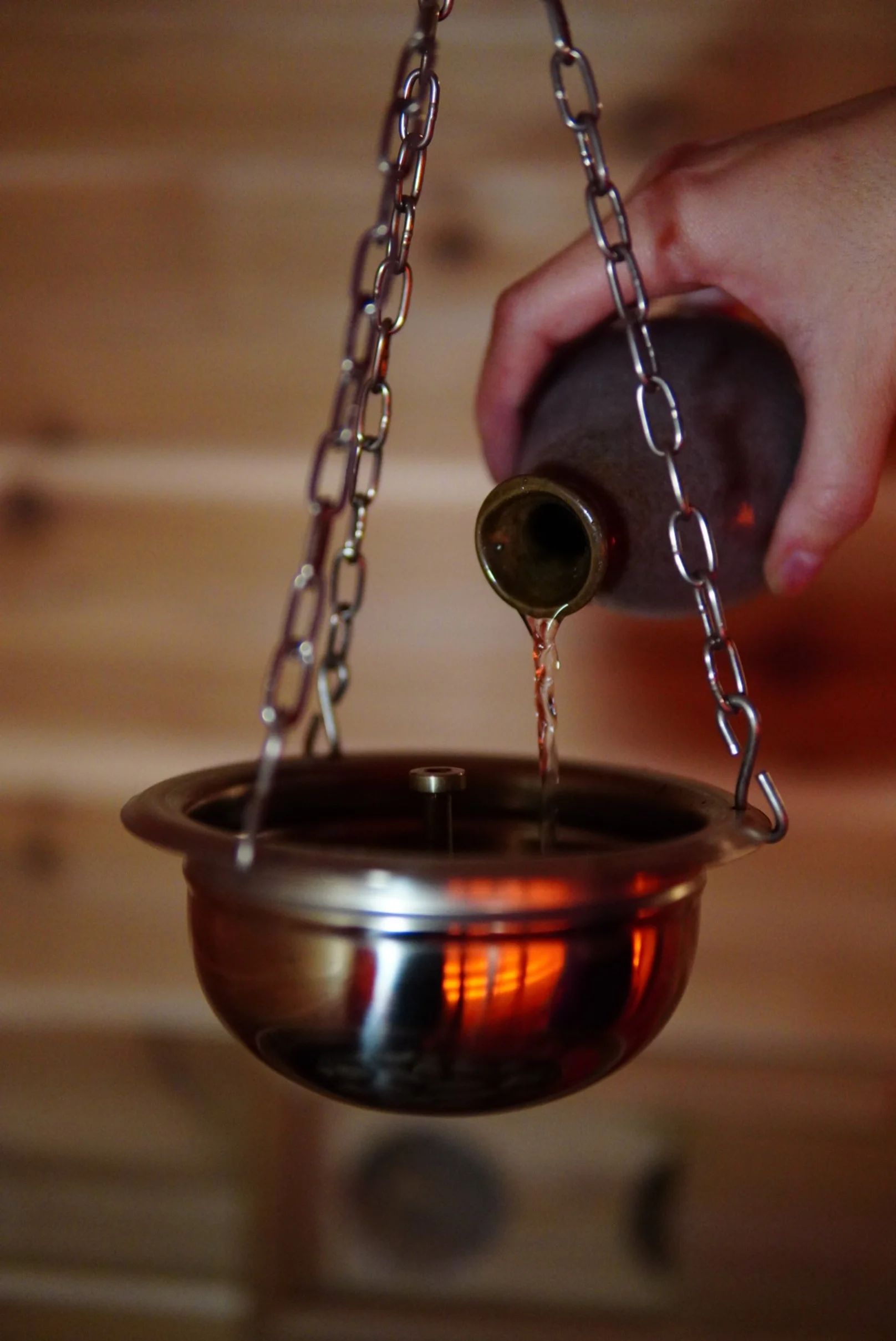 Close-up of a hand pouring liquid from a bottle into a brass bowl suspended from a chain, with a wooden background.