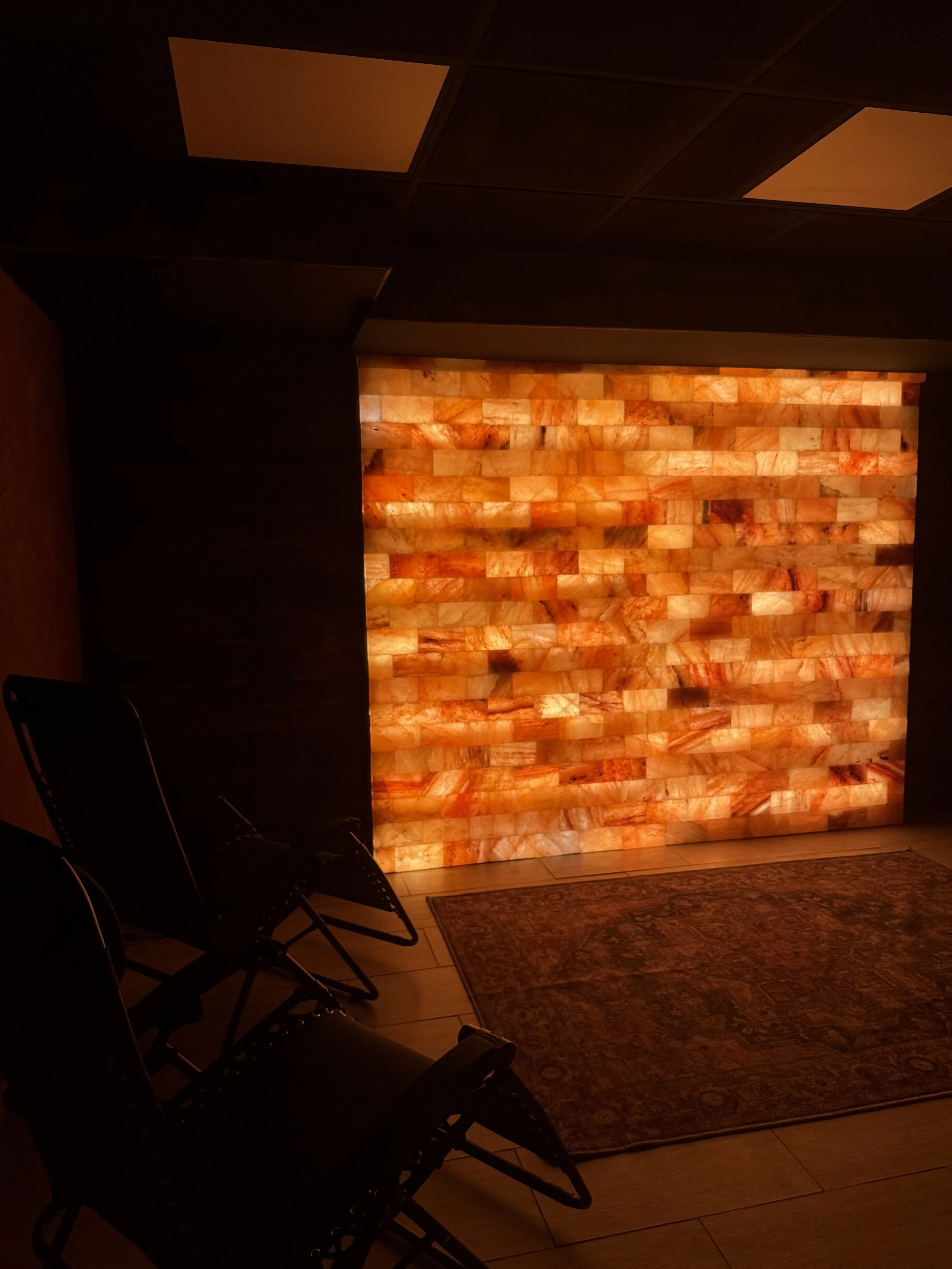 A dimly lit room with a salt rock wall illuminated from behind, creating a warm glow. There are a few black chairs and a patterned rug on the hardwood floor.