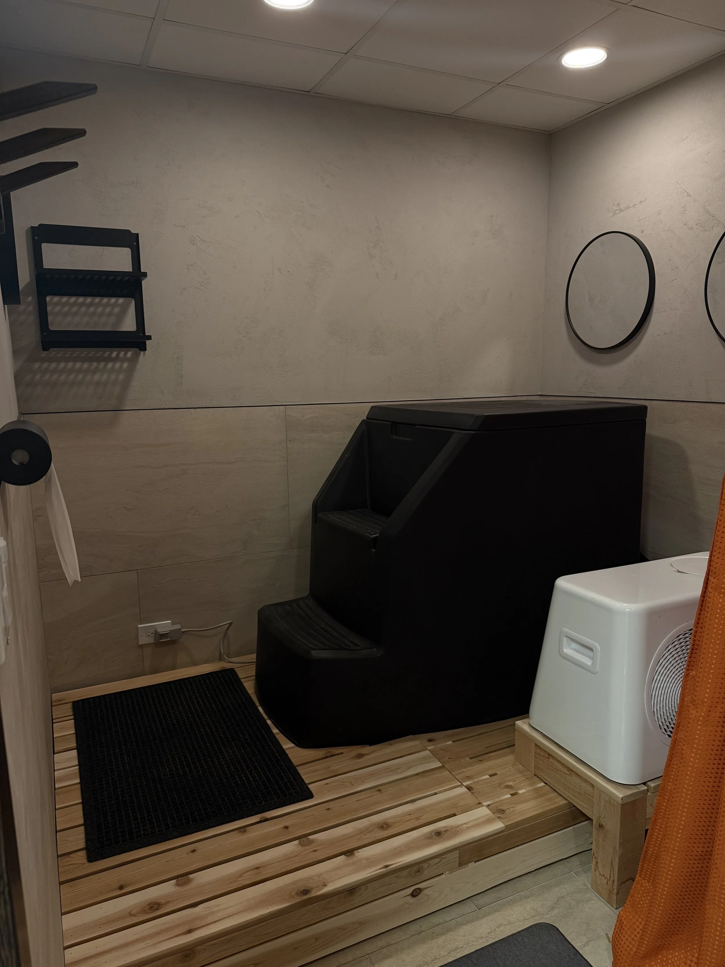 Small room with beige walls and ceiling, featuring a black foot bath, a white appliance, mirror, black shelf, black mat, and orange curtain.