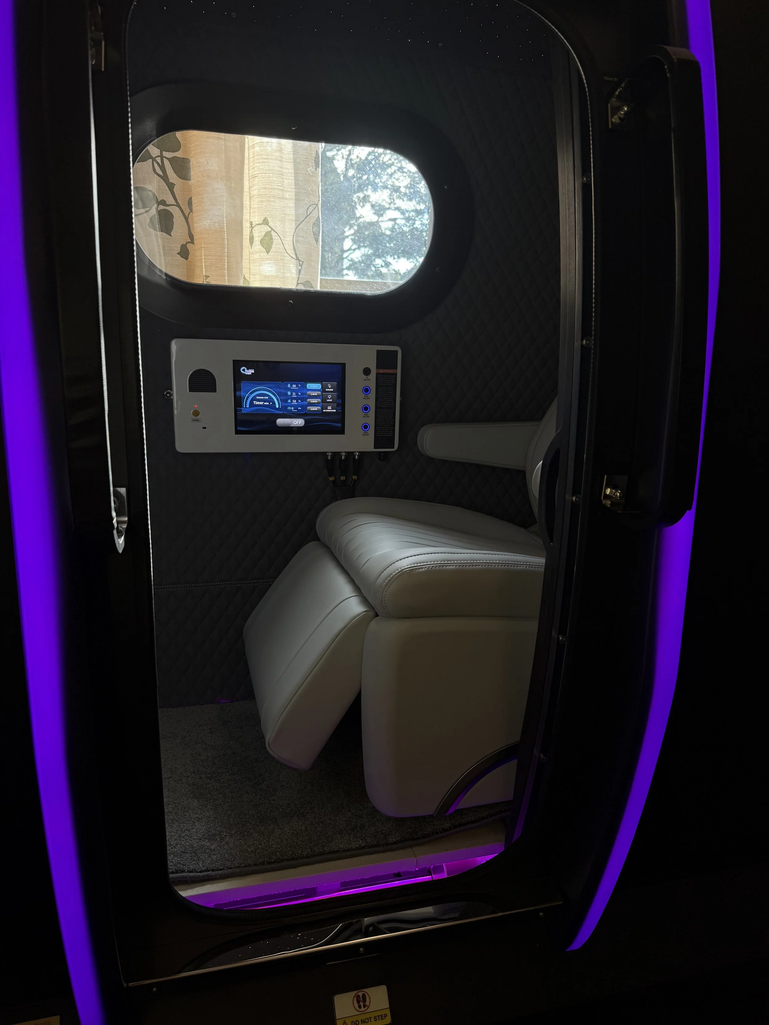 Inside a futuristic pod or small room with a window showing trees, containing a cushioned white chair and a control panel with a digital screen.