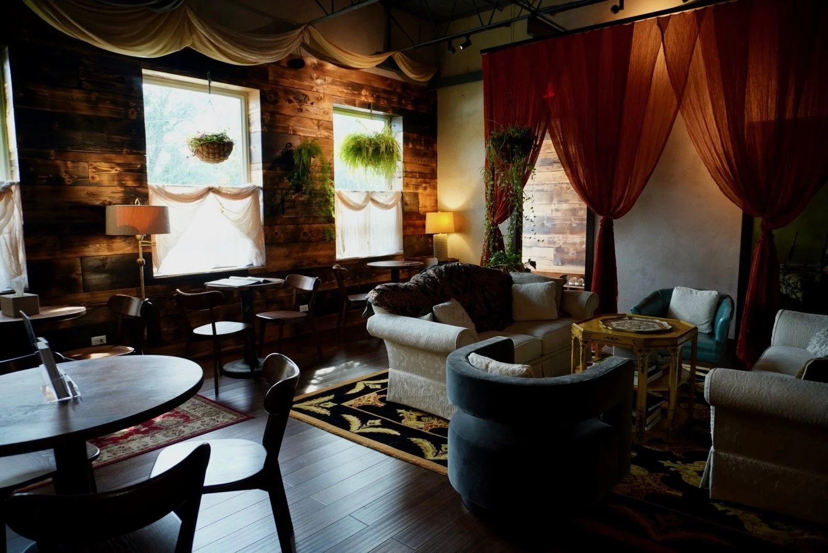 A cozy living room with wooden walls, large windows with white curtains, and various seating including sofas and chairs. There are plants hanging by the windows, lamps providing warm lighting, and a decorative area rug on the wooden floor. Red curtains separate part of the room.