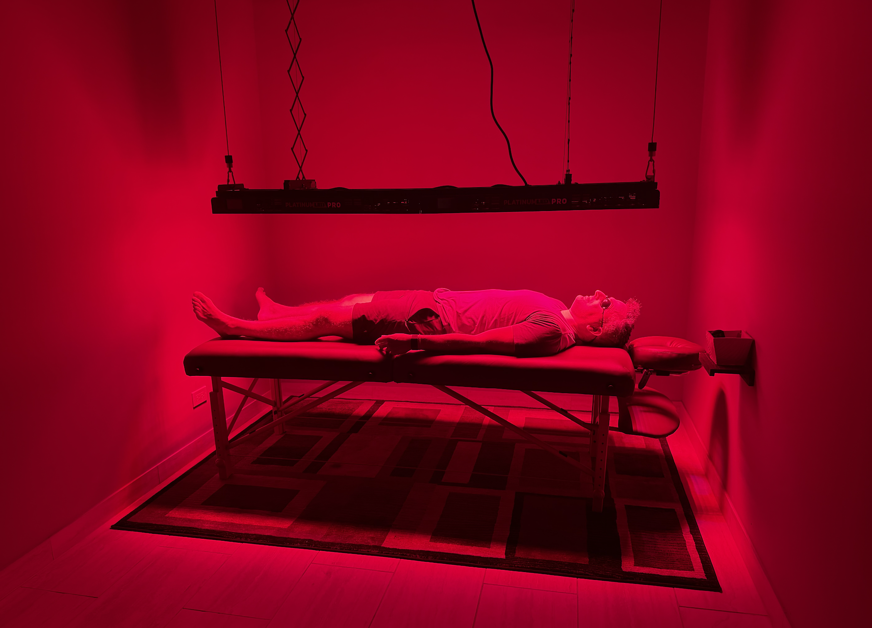 A man lying on a treatment table inside a red-lit room, wearing eye protection, with a hanging LED grow light above him. The room has a patterned rug on the floor and a small stool next to the treatment table.