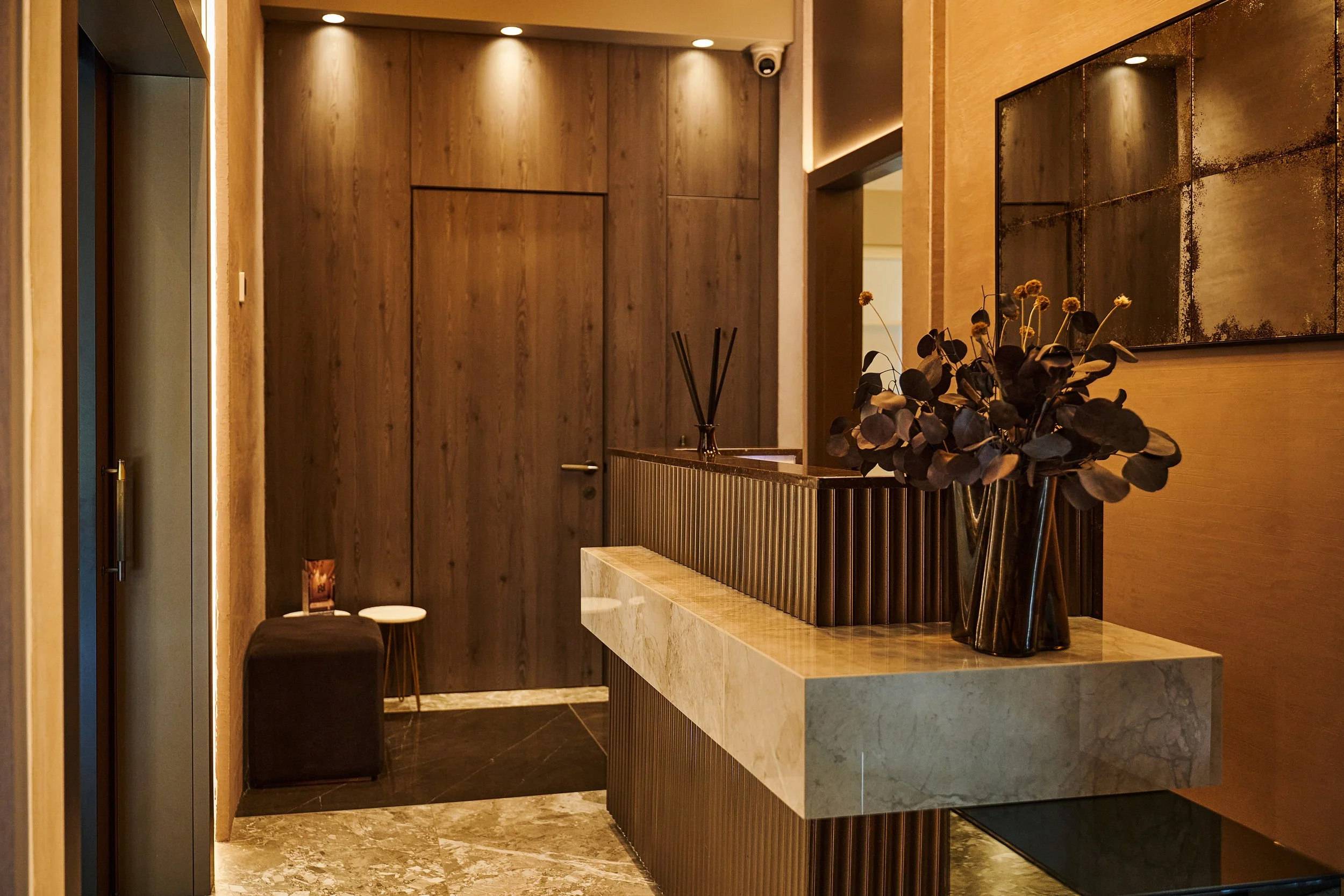 Interior of a modern, warmly lit lobby or reception area featuring a wood-paneled wall, a marble-topped counter with a dark decorative panel, a vase of dried flowers, and a large abstract painting on the wall.