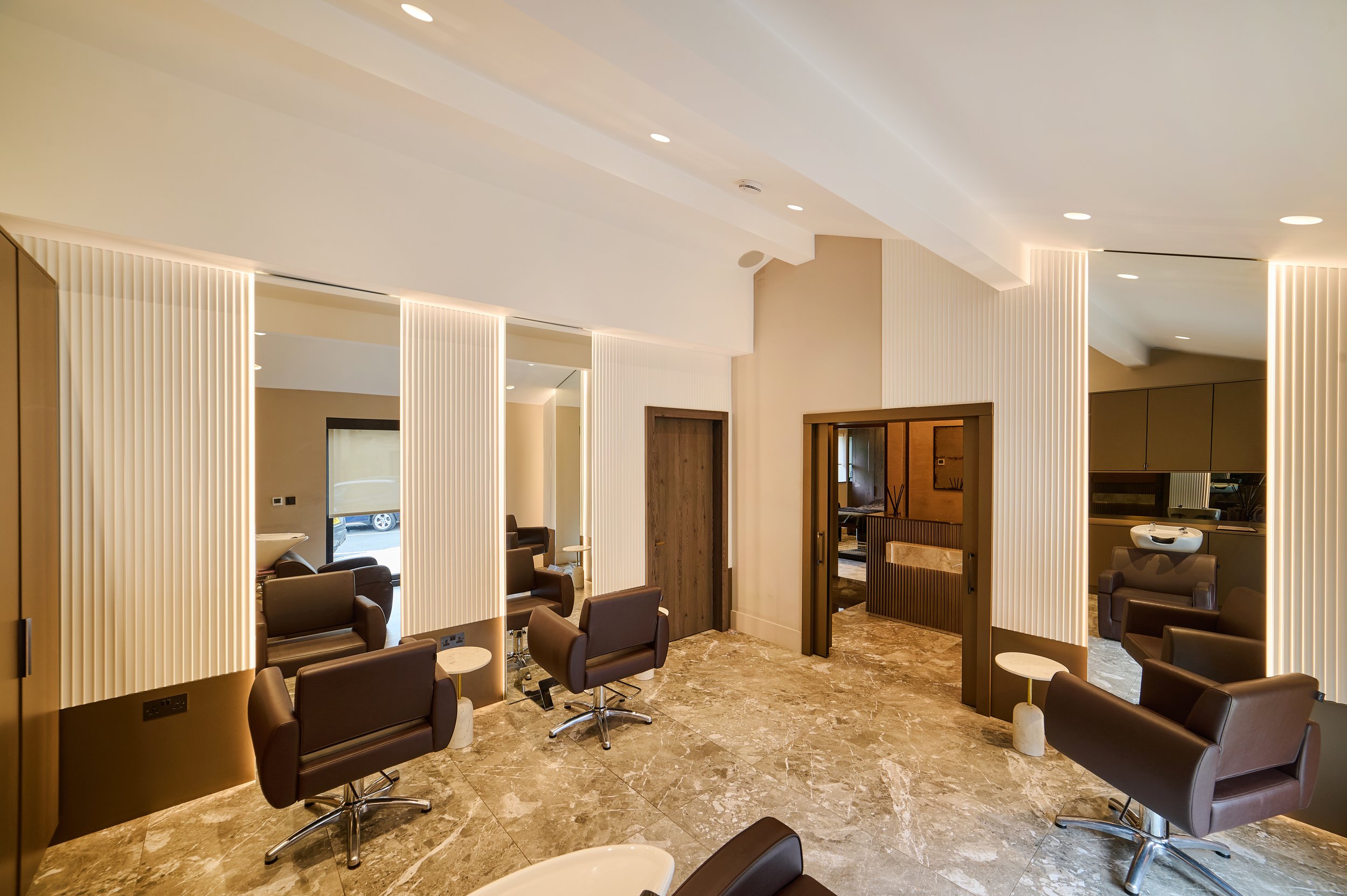 Modern salon with brown chairs, mirrors, marble floor, and beige walls illuminated by spotlights.