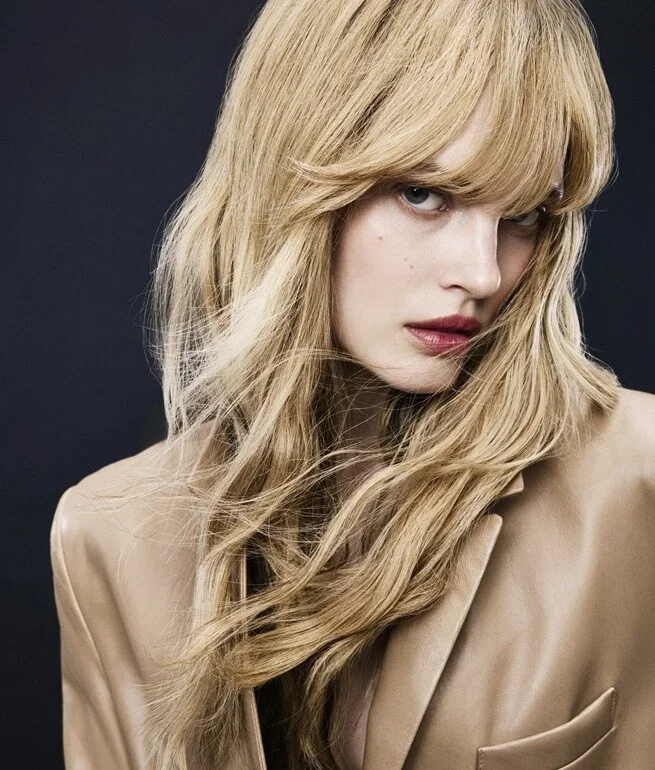Close-up portrait of a woman with long blonde wavy hair partially covering her face, wearing a beige blazer against a dark background.