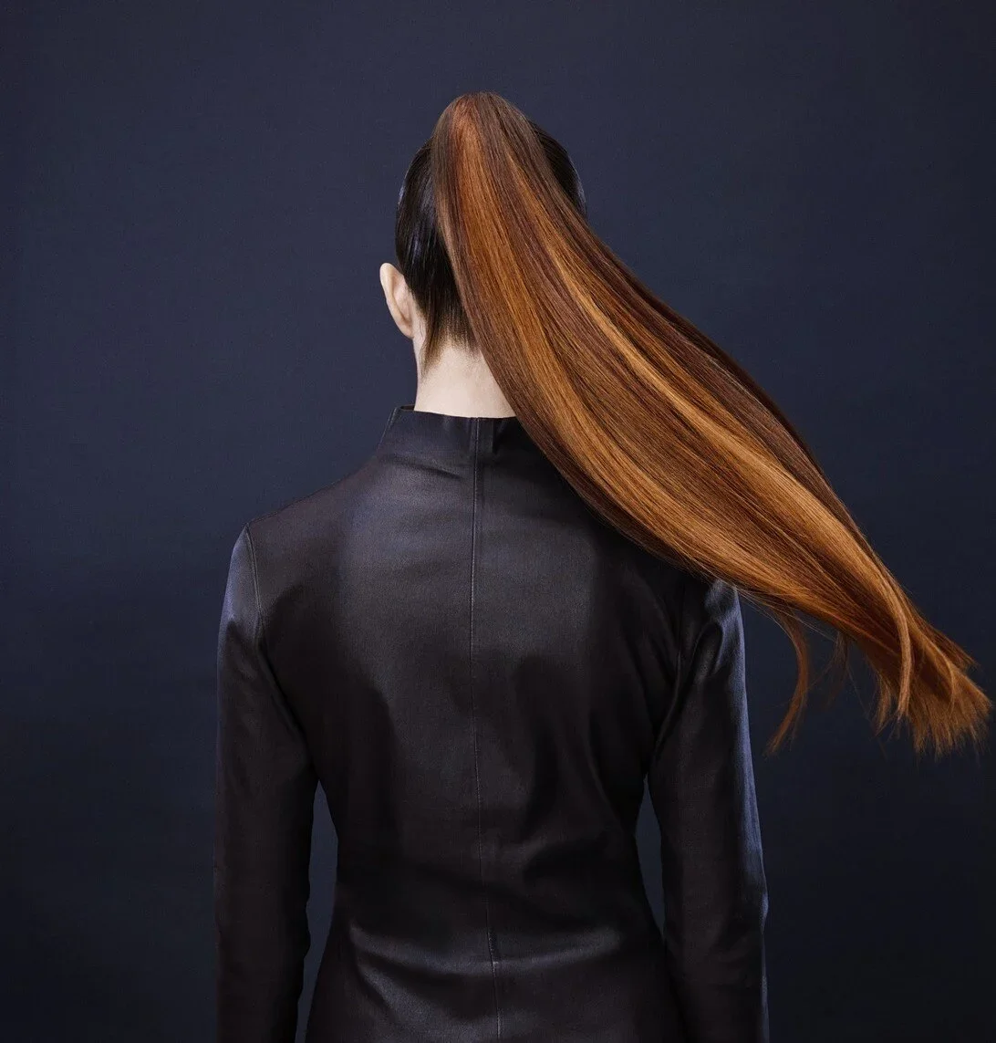 Woman with long, straight, reddish-brown hair, wearing a black jacket, facing away against a dark background.