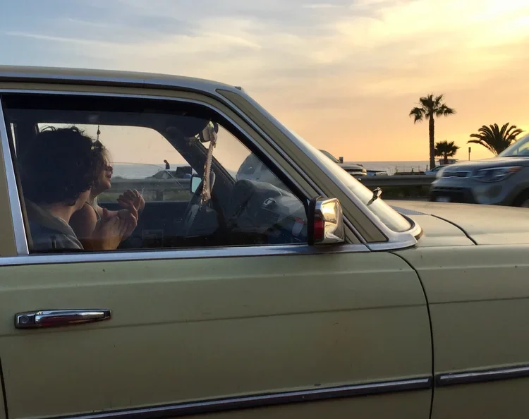 Person sitting inside a yellow car, holding a sandwich, with a coastal sunset in the background. Palm trees and a road with cars are visible outside.