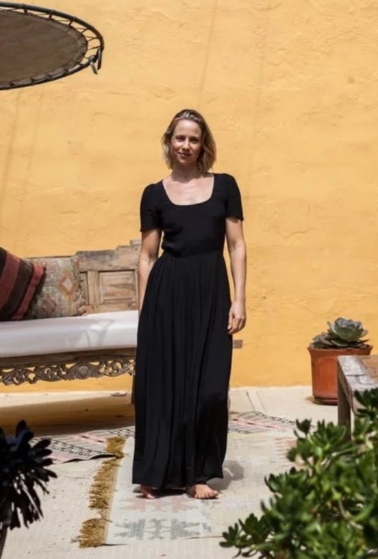A woman in a black dress standing barefoot on a patterned outdoor rug in front of a yellow wall, with a wooden bench, cushions, and potted plants around her.