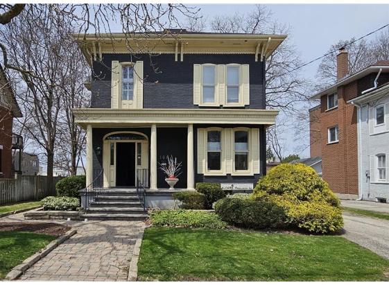 Front view of a two-story house with black siding, cream trim, and a front porch with steps, surrounded by trees and landscaping.