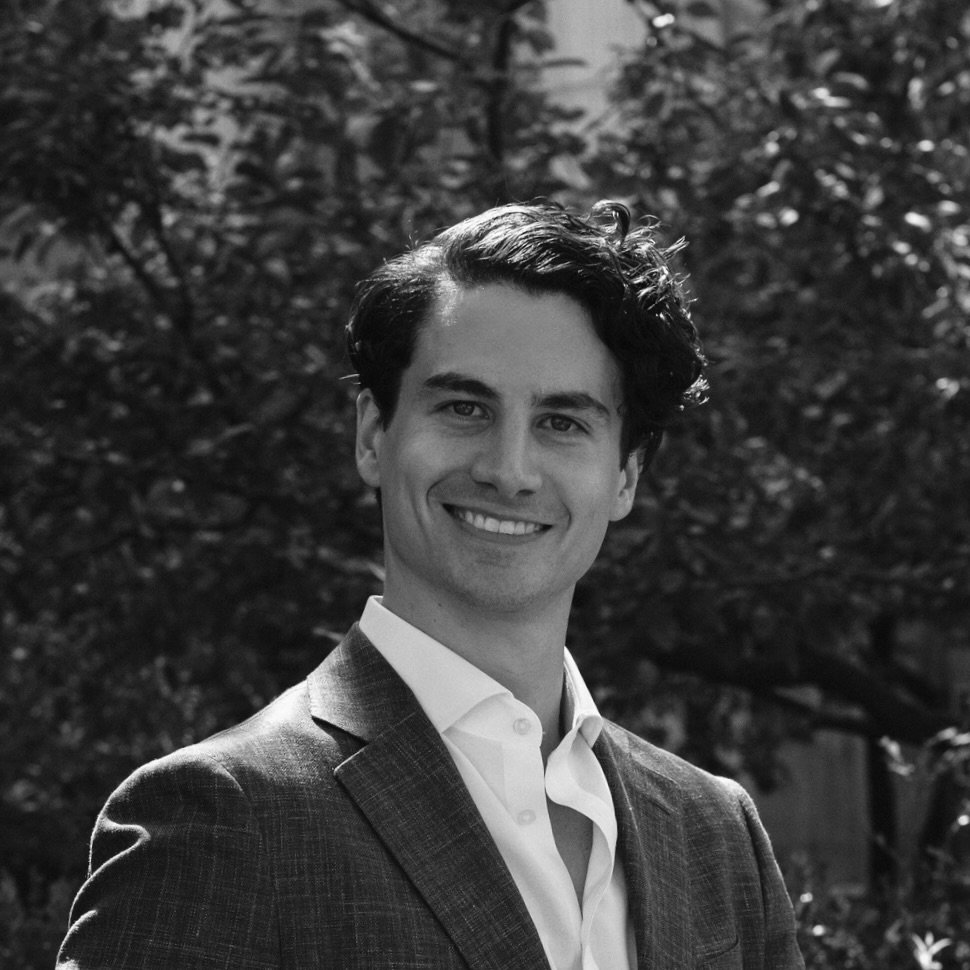 A young man in a suit smiling outdoors with trees in the background.