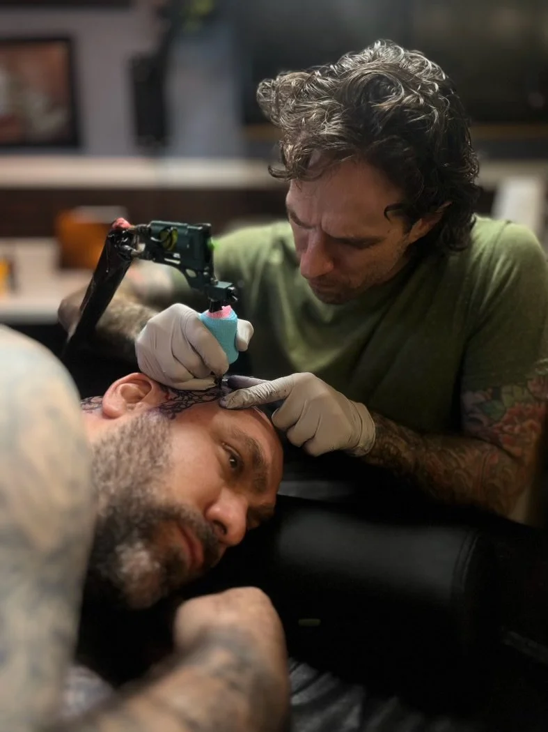An artist with curly hair and tattoos is tattooing a man's forehead in a studio, using a tattoo machine and wearing gloves.