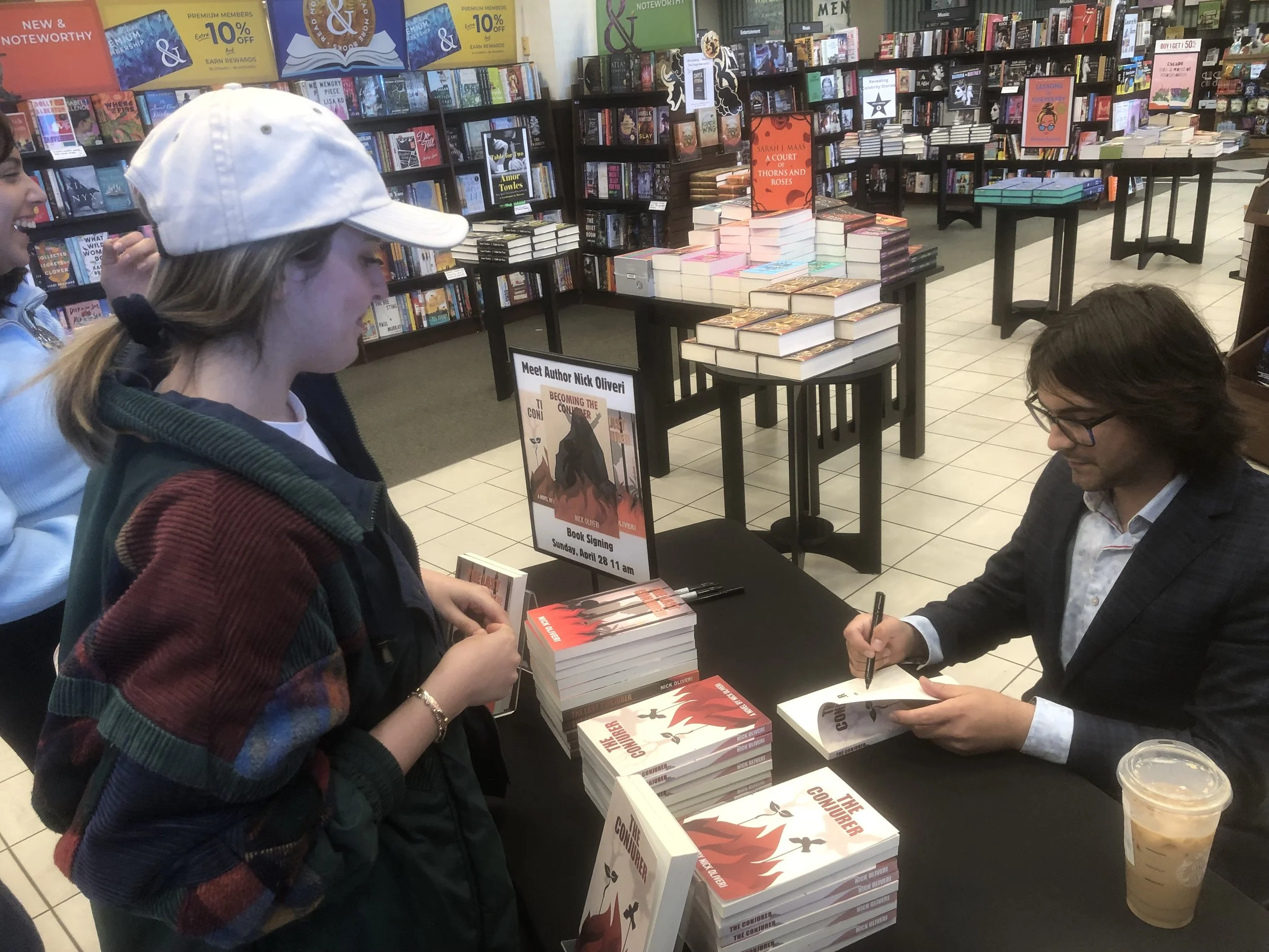 A bookstore with tables of stacked books, people waiting for a book signing event, and posters advertising the author Nick Oliveri's signing of his book 'The Conqueror.'
