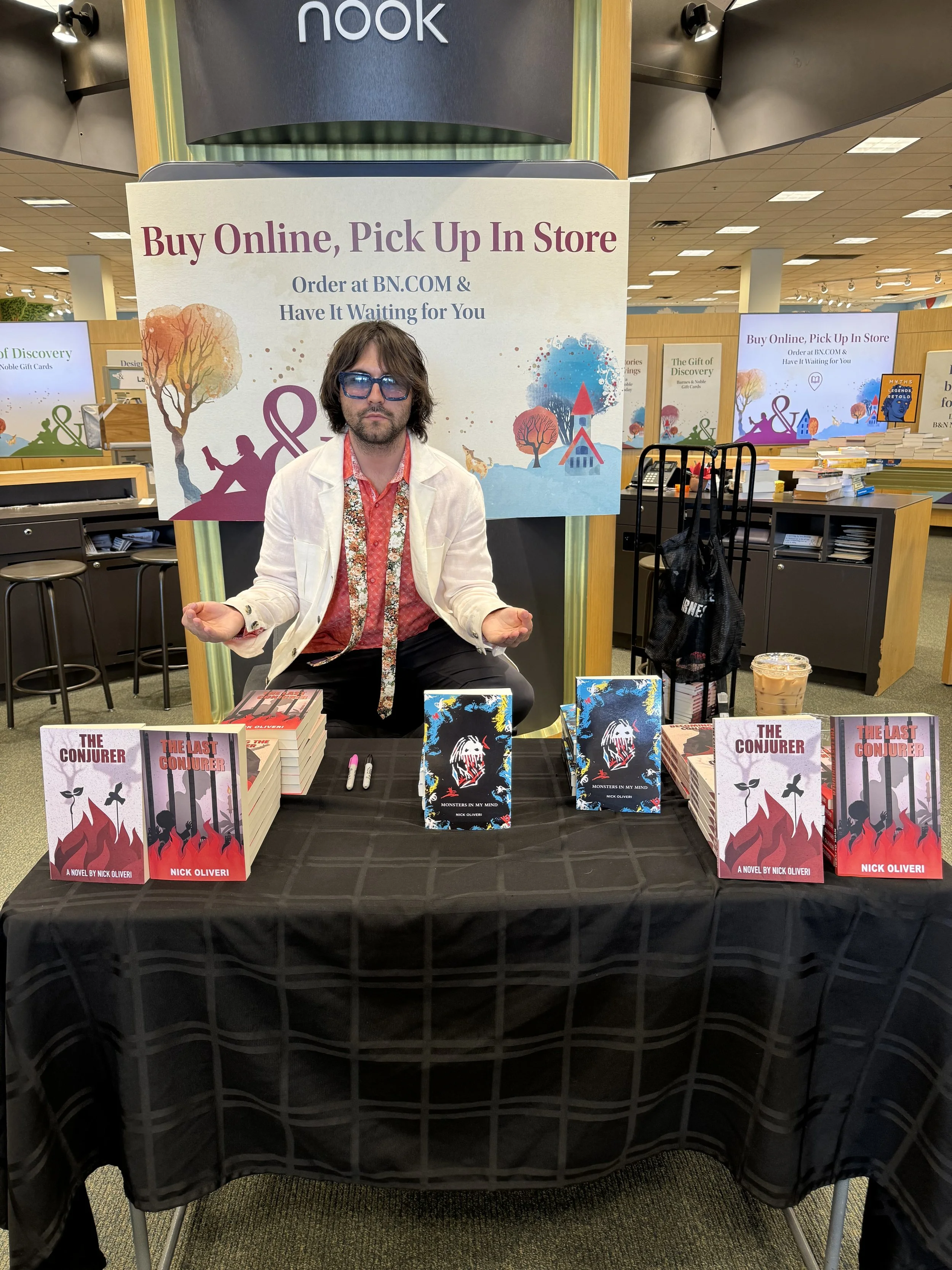 A man sitting at a book signing table in a bookstore or library. The table displays copies of the book titled 'The Conjurer' by Nick Oliveri and promotional materials. There are bookshelves and a large sign behind him that says 'Buy Online, Pick Up In Store' with additional text about ordering at BN.COM.