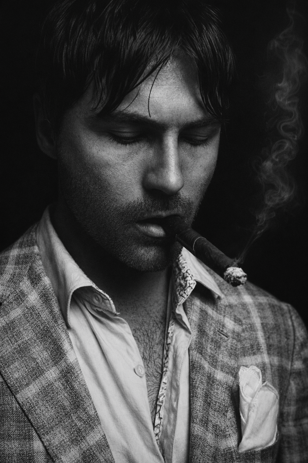 A black and white photo of a man with closed eyes, wearing a suit jacket and a dress shirt, smoking a cigar with smoke curling up beside his face.