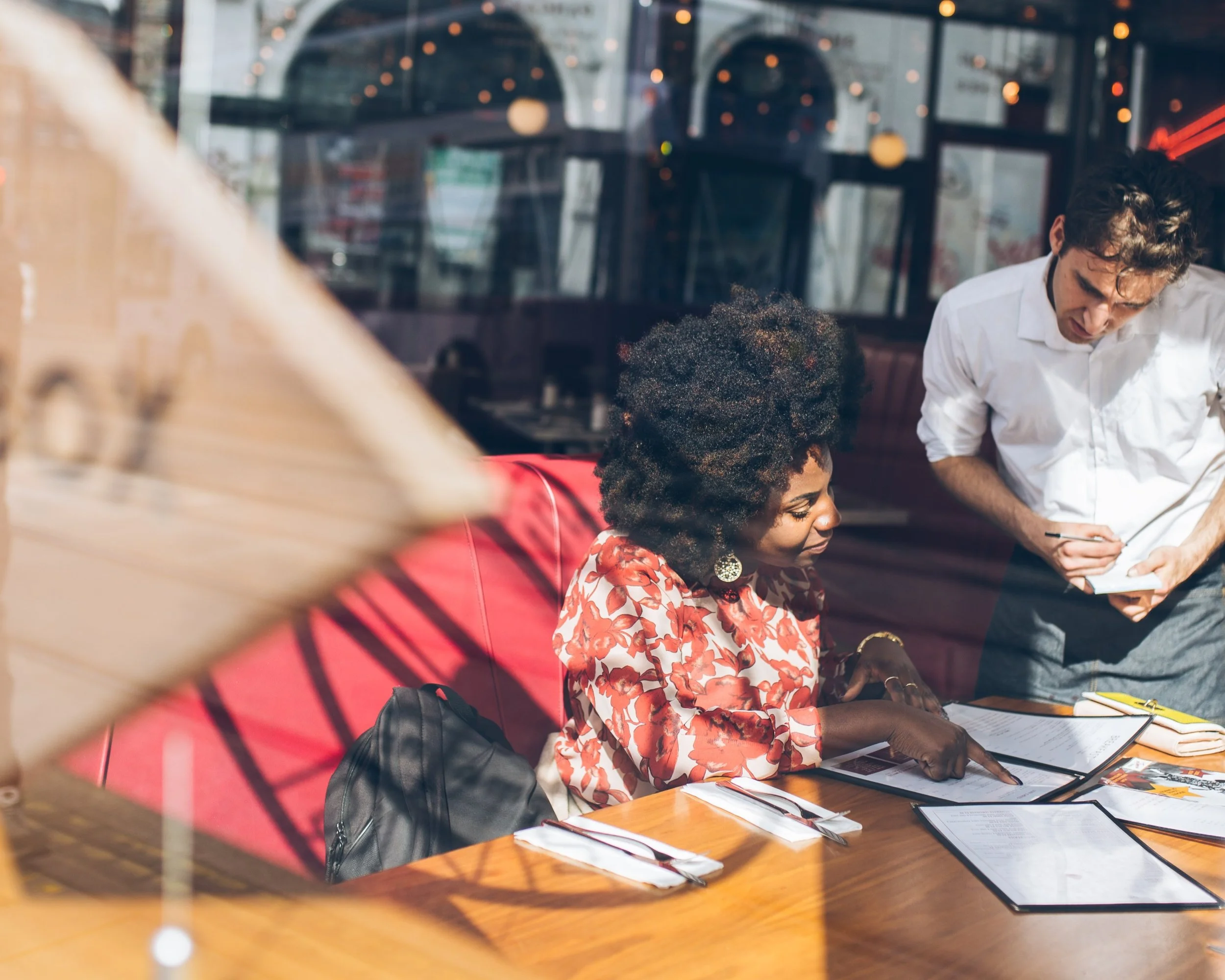 A woman with curly black hair wearing a red and white floral top is sitting at a restaurant table with menus, pointing at a menu. A man with dark curly hair in a white shirt is standing next to her, taking notes on a notepad.