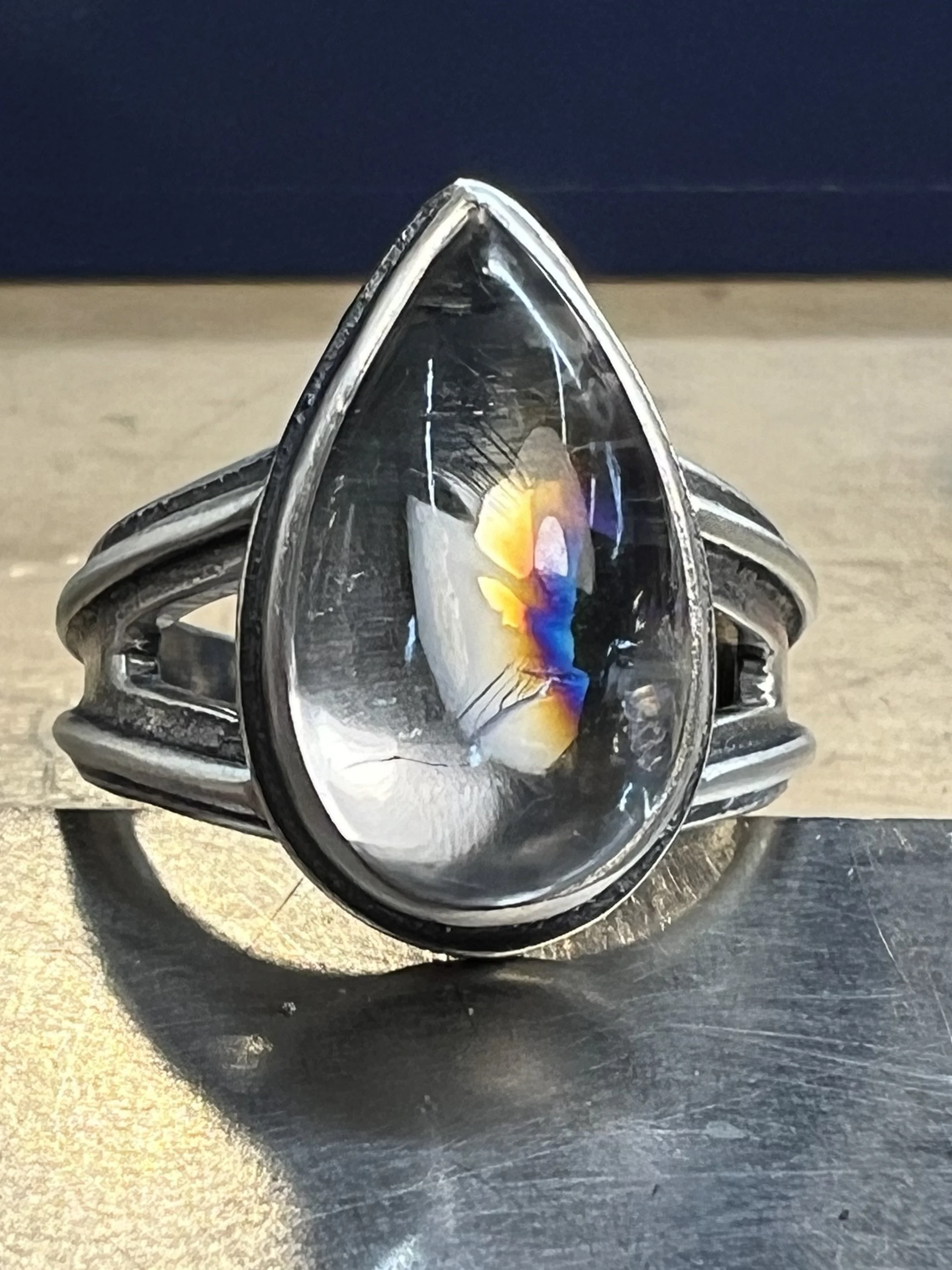 Close-up of a sterling silver ring with a large, teardrop crystal and a colorful rainbow reflection inside the gemstone, resting on a textured surface.