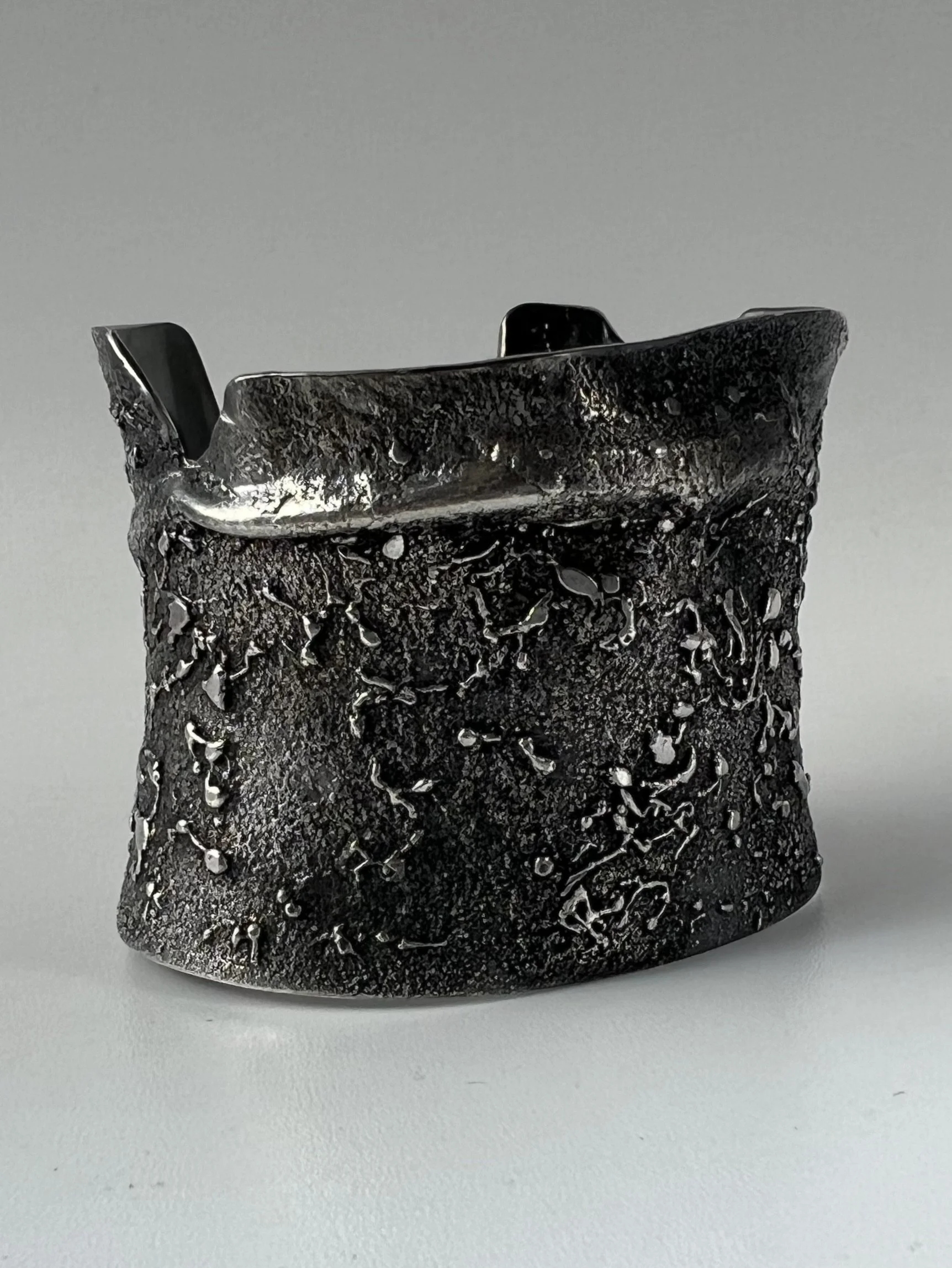 Close-up of oxidized sterling silver cuff with a textured surface, placed on a plain light gray background.