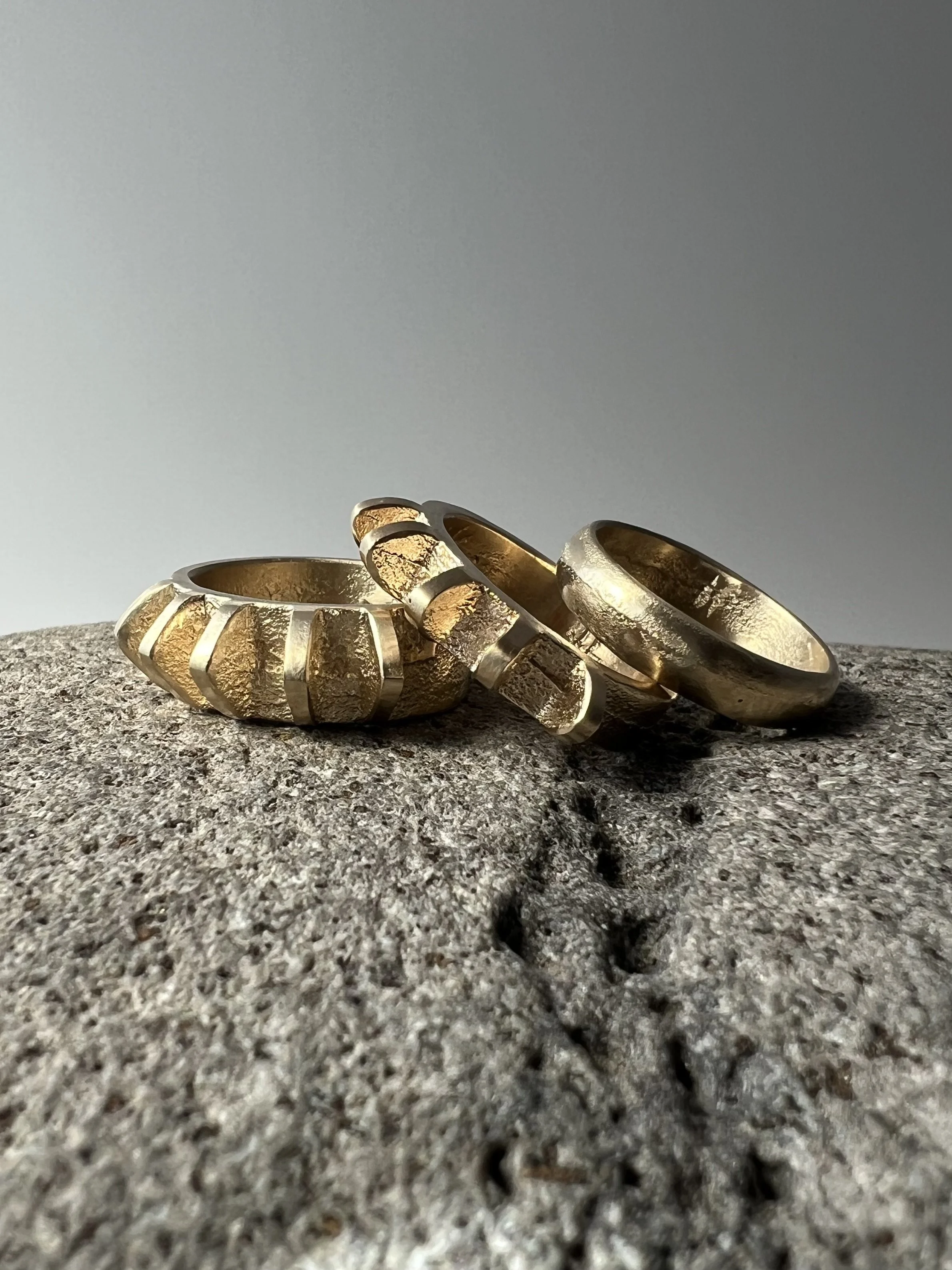 Three gold rings with textured and polished surfaces resting on grey textured surface, shadow cast on ground.