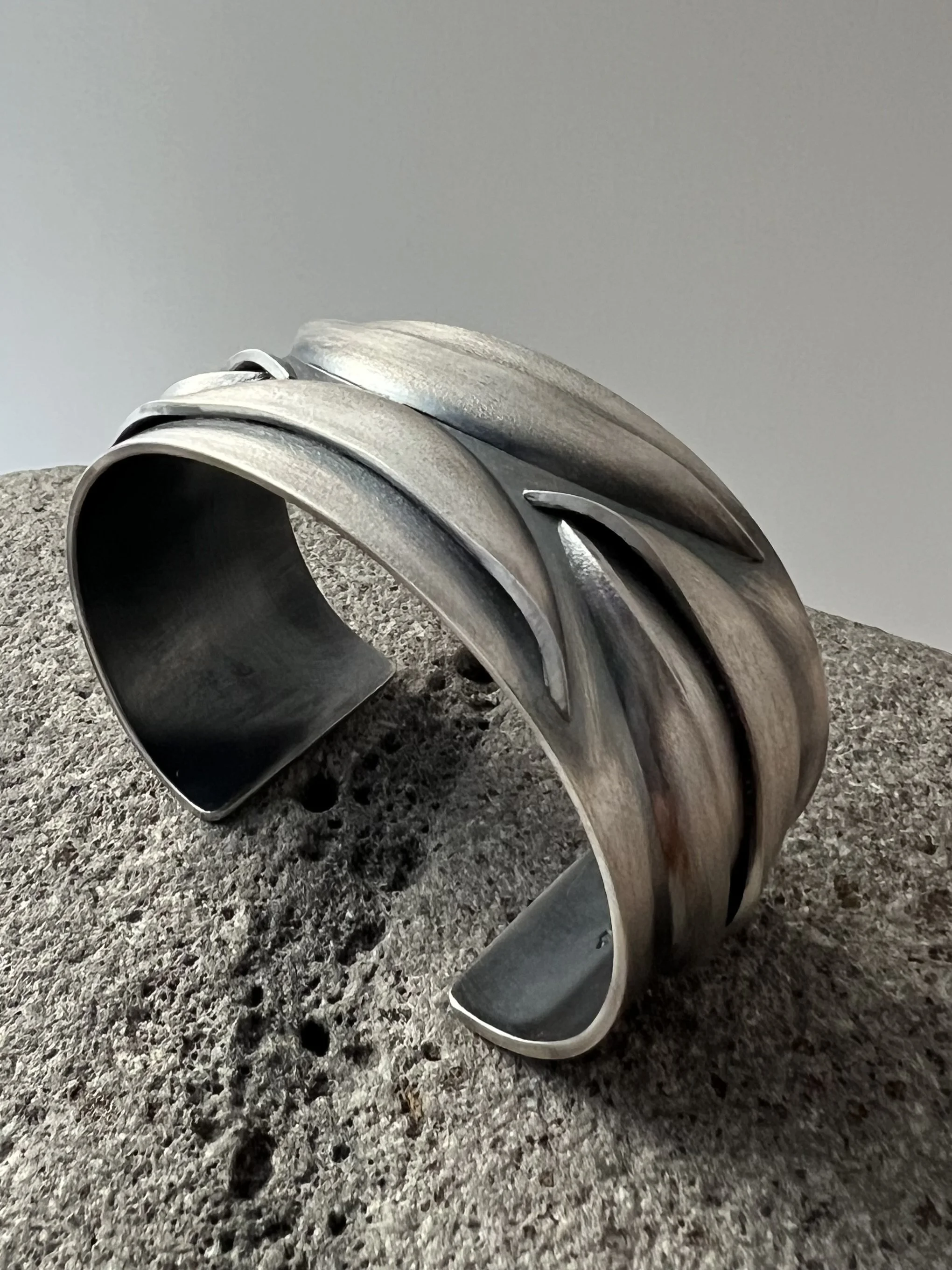 Close-up of a sterling silver cuff with multiple curved, elongated, and overlapping components placed on a rough gray surface against a plain light background.