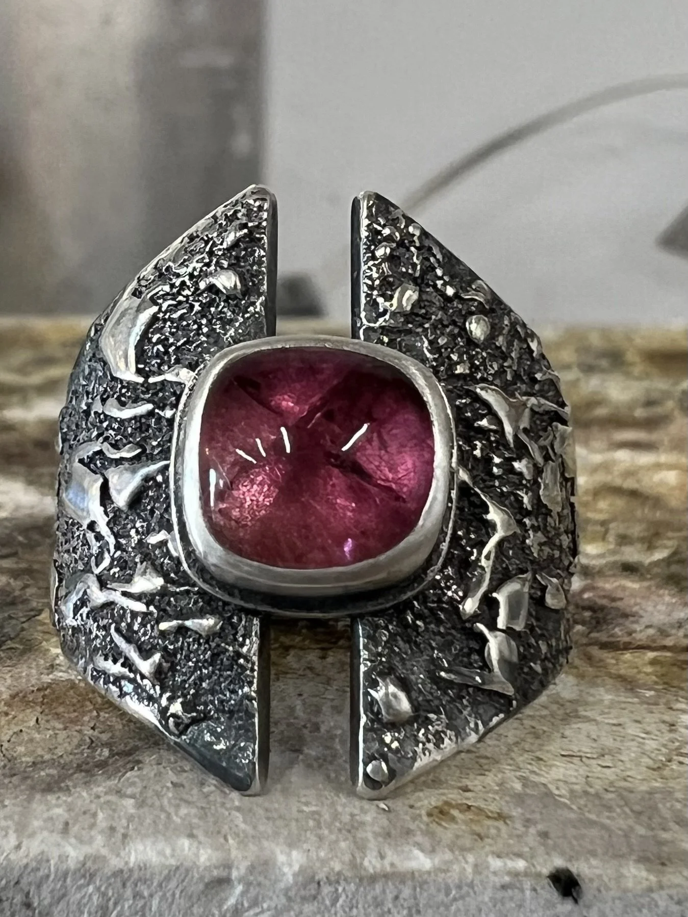 A sterling silver ring with a large red gemstone set in the center, surrounded by textured silver details resembling wings, placed on a wooden surface.