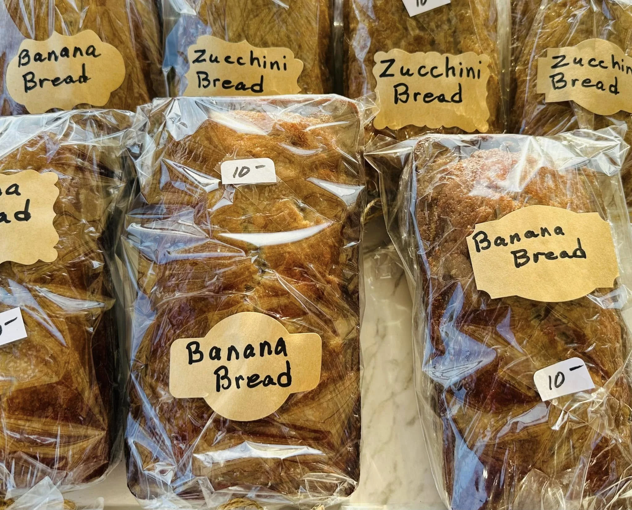 Multiple loaves of zucchini bread wrapped in plastic and labeled with handwritten signs that say 'Zucchini Bread'. The loaves are on display, some with price tags.