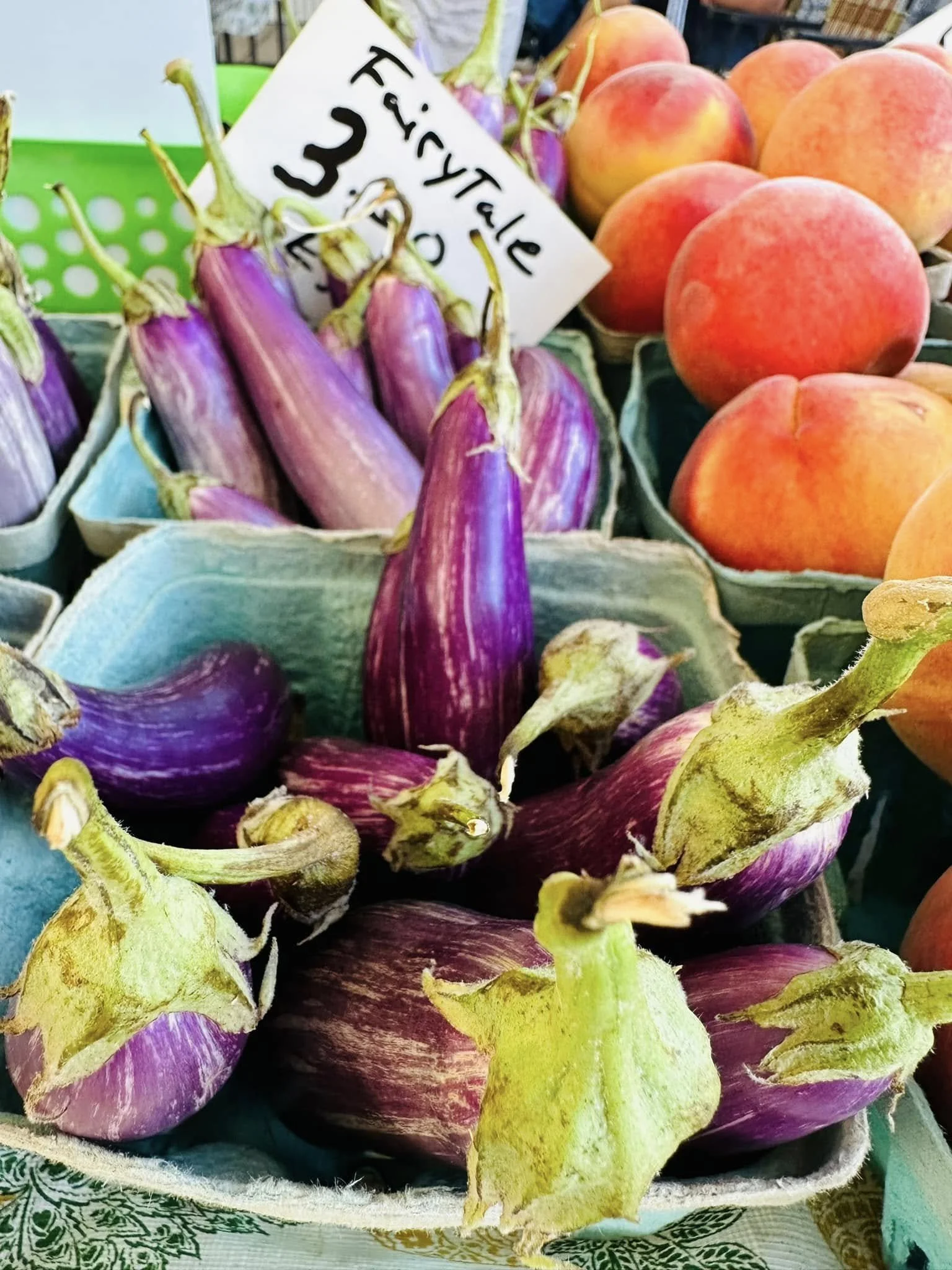 A basket of fresh peaches and purple eggplants with a handwritten price sign at a market.