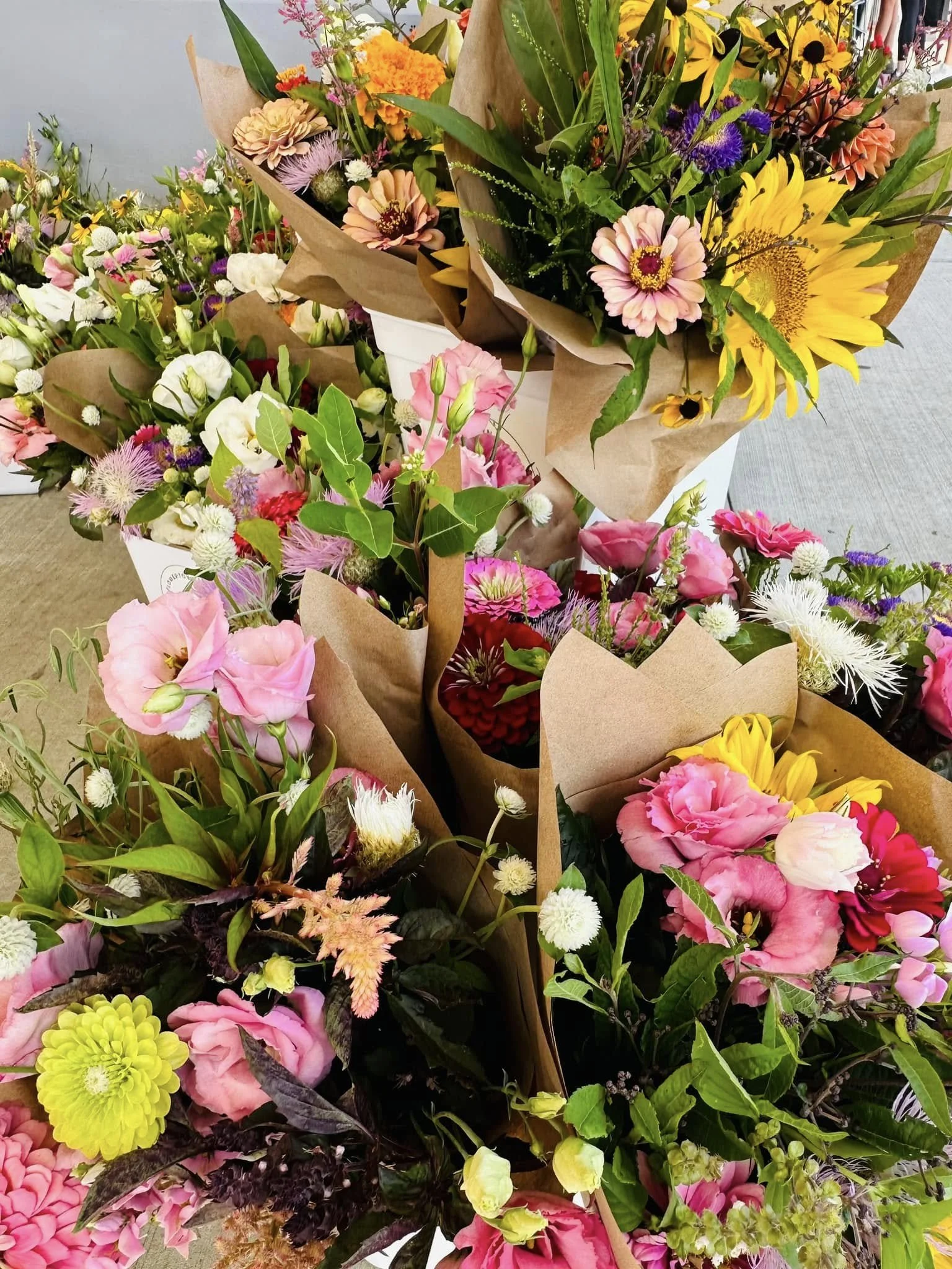 A variety of colorful flowers arranged in bouquets covered with brown craft paper, including sunflowers, pink roses, white and purple blooms, and green leaves.