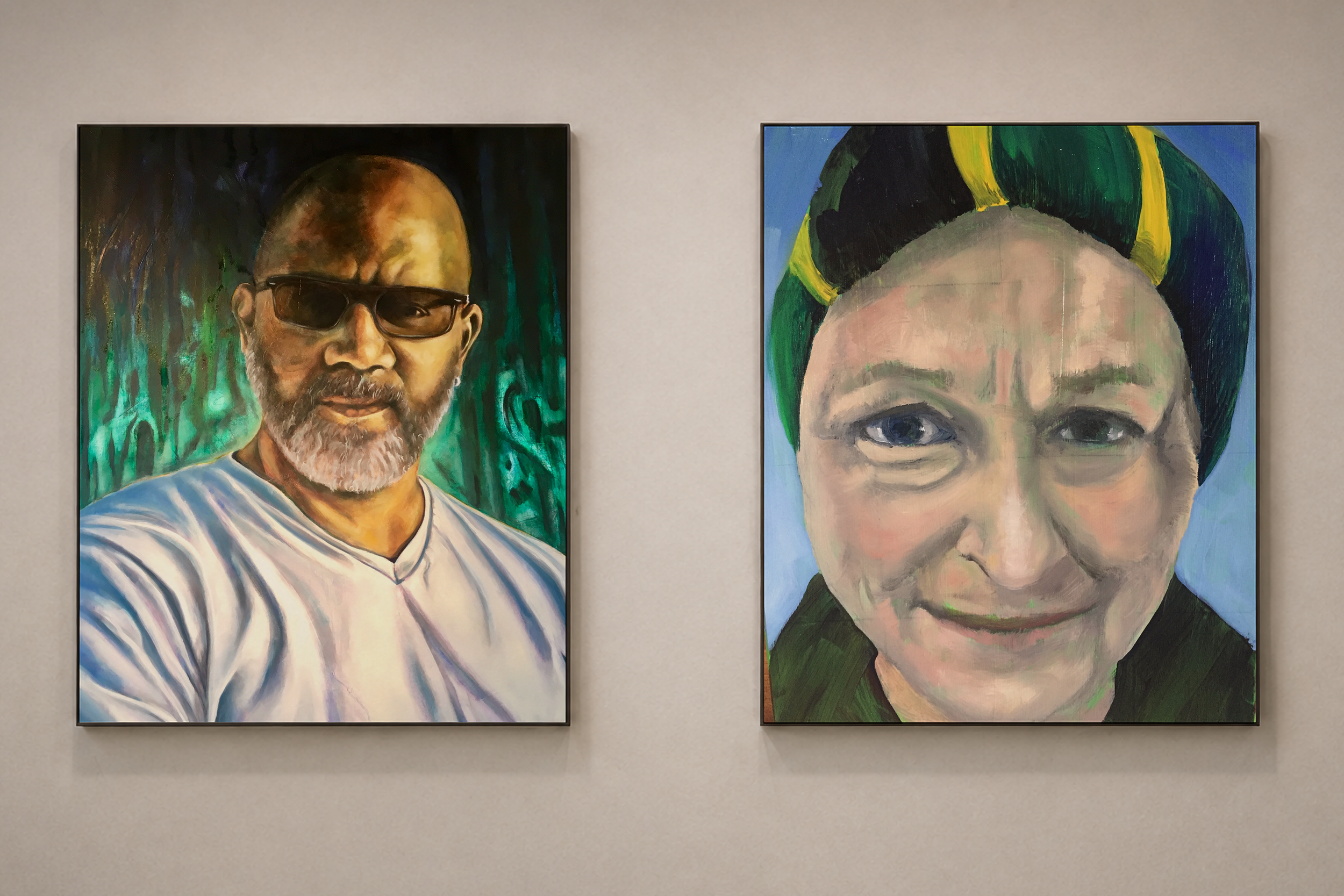Two paintings of people hanging on a wall. The left painting depicts a man with a beard, glasses, and a striped shirt, with a colorful background. The right painting depicts a face of a person with short hair and an expressive face, with a blue backg