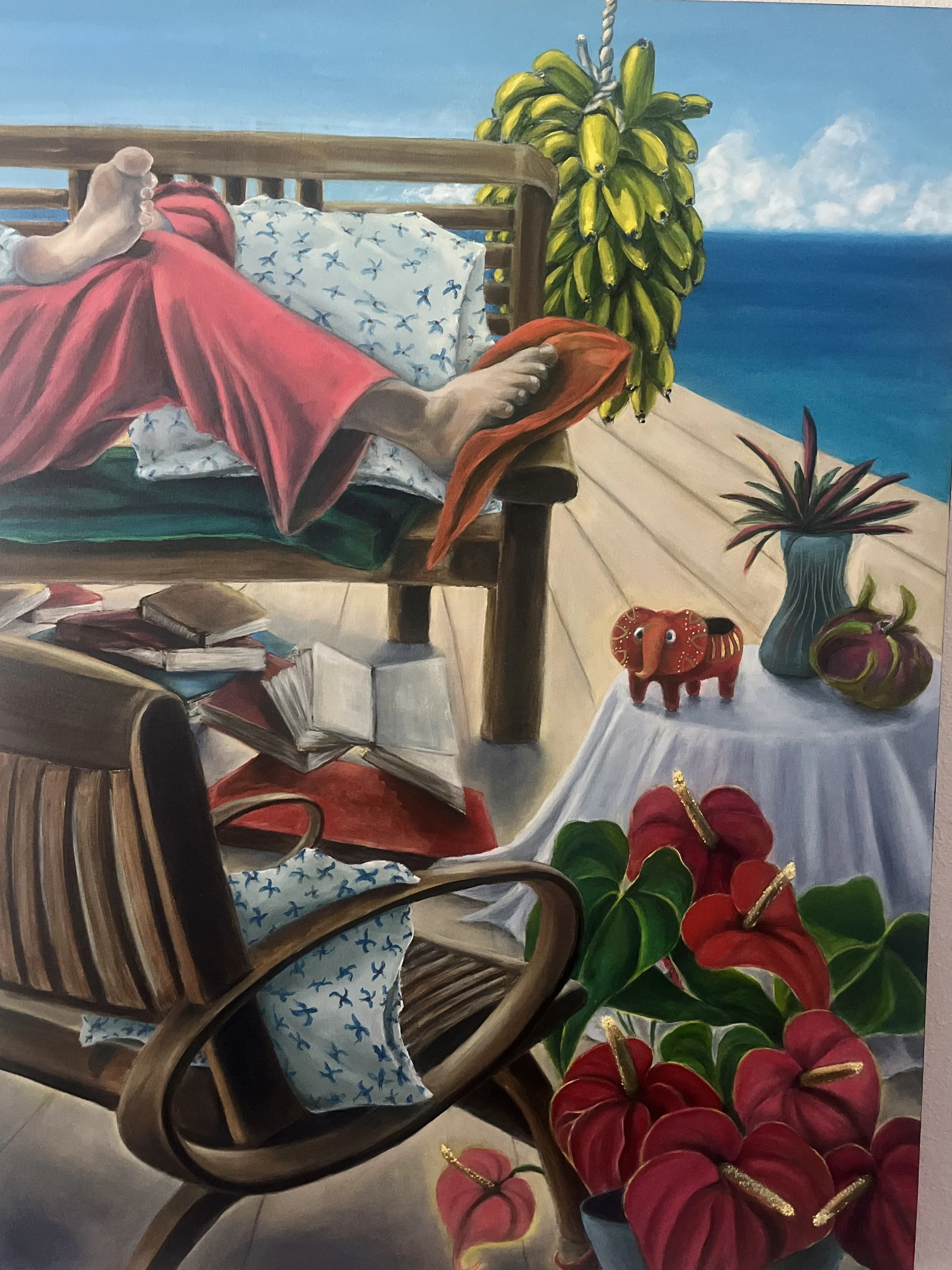 A painting of a person lying in a wooden chair on a deck, with feet visible. The person is wearing a red skirt and a white blouse with blue floral pattern. There are multiple books on the floor near the chair, and a basket of red flowers nearby. A bunch of bananas hangs in the background against a blue sky with some clouds and the ocean.