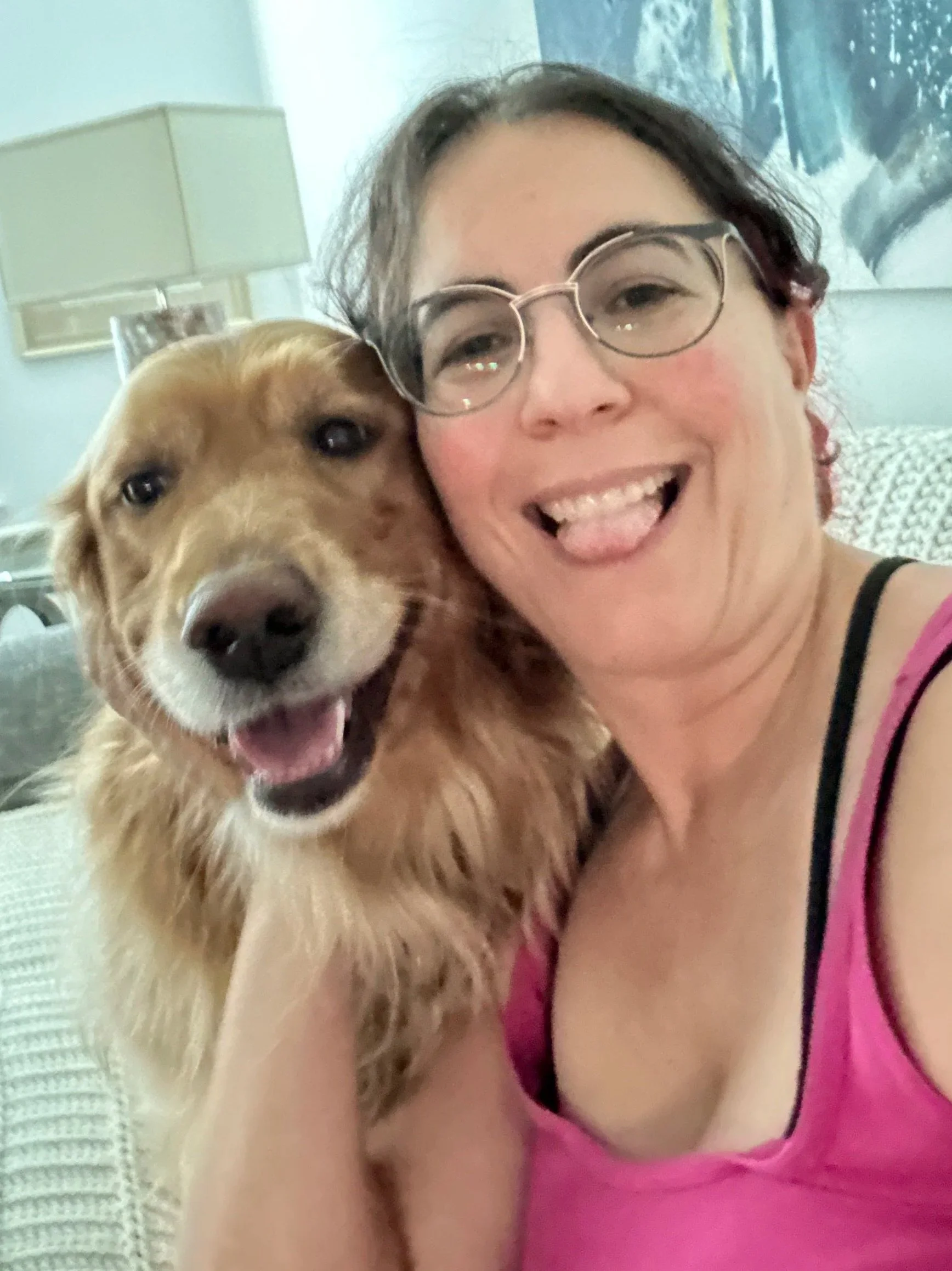 A woman with glasses and a pink tank top posing with her mouth open and tongue out, hugging a happy golden retriever dog with its tongue out, in a room with a lamp and a framed picture in the background.