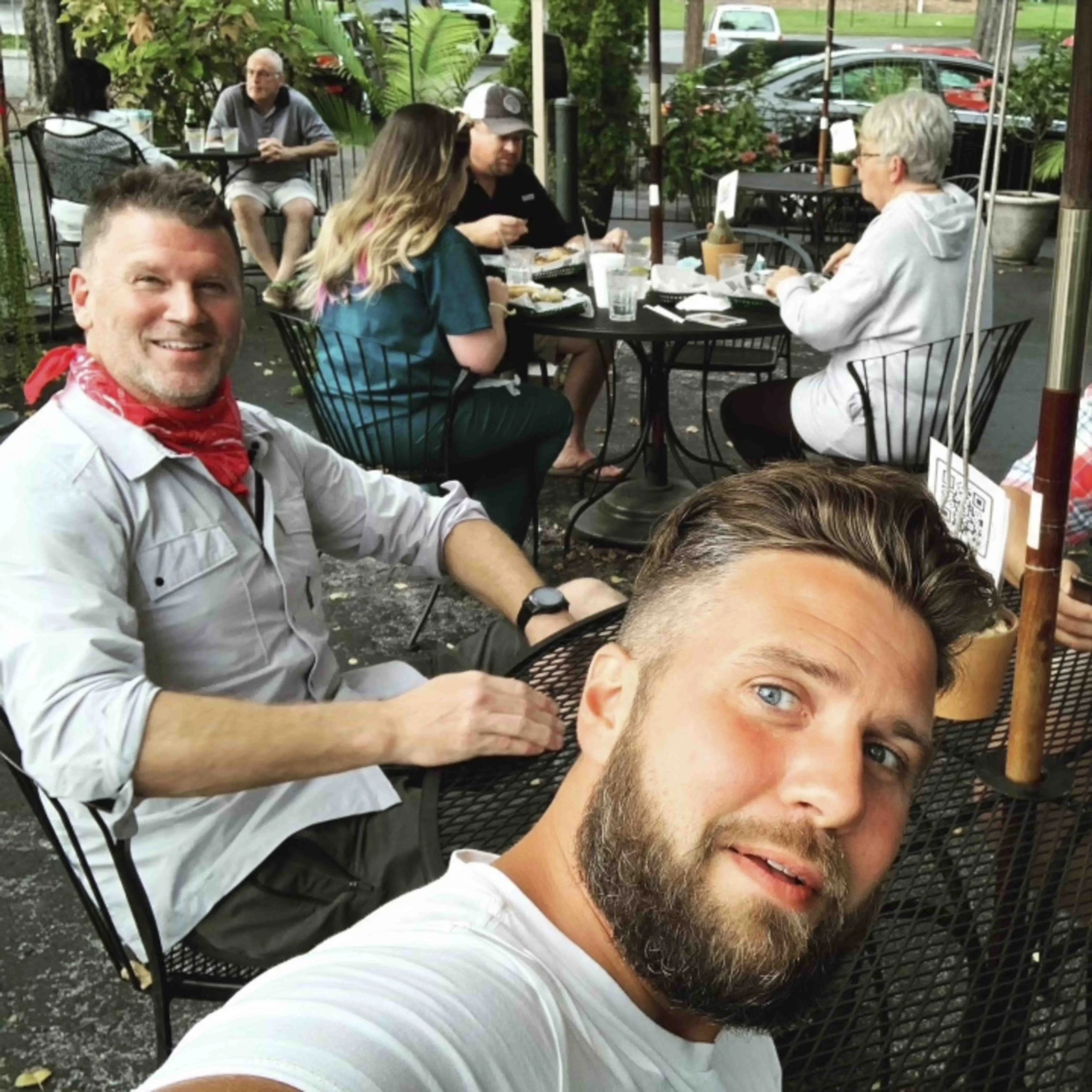 A group of people sitting and eating at a black metal outdoor patio table, with lush green plants and parked cars in the background.