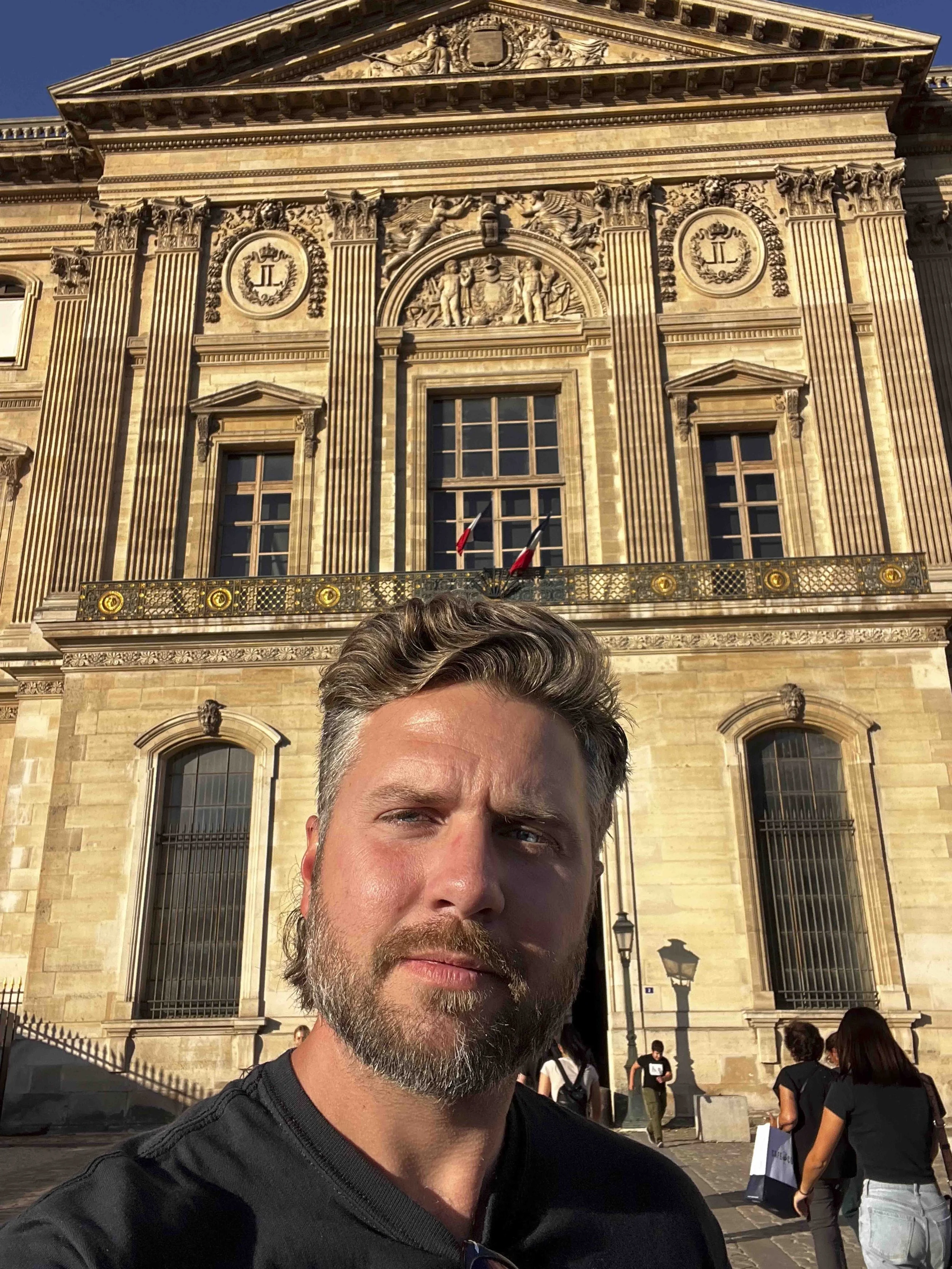 A man with a beard and brown hair taking a selfie in front of an ornate historic building with classical architecture, detailed sculptures, and large windows, with a group of people in the background.