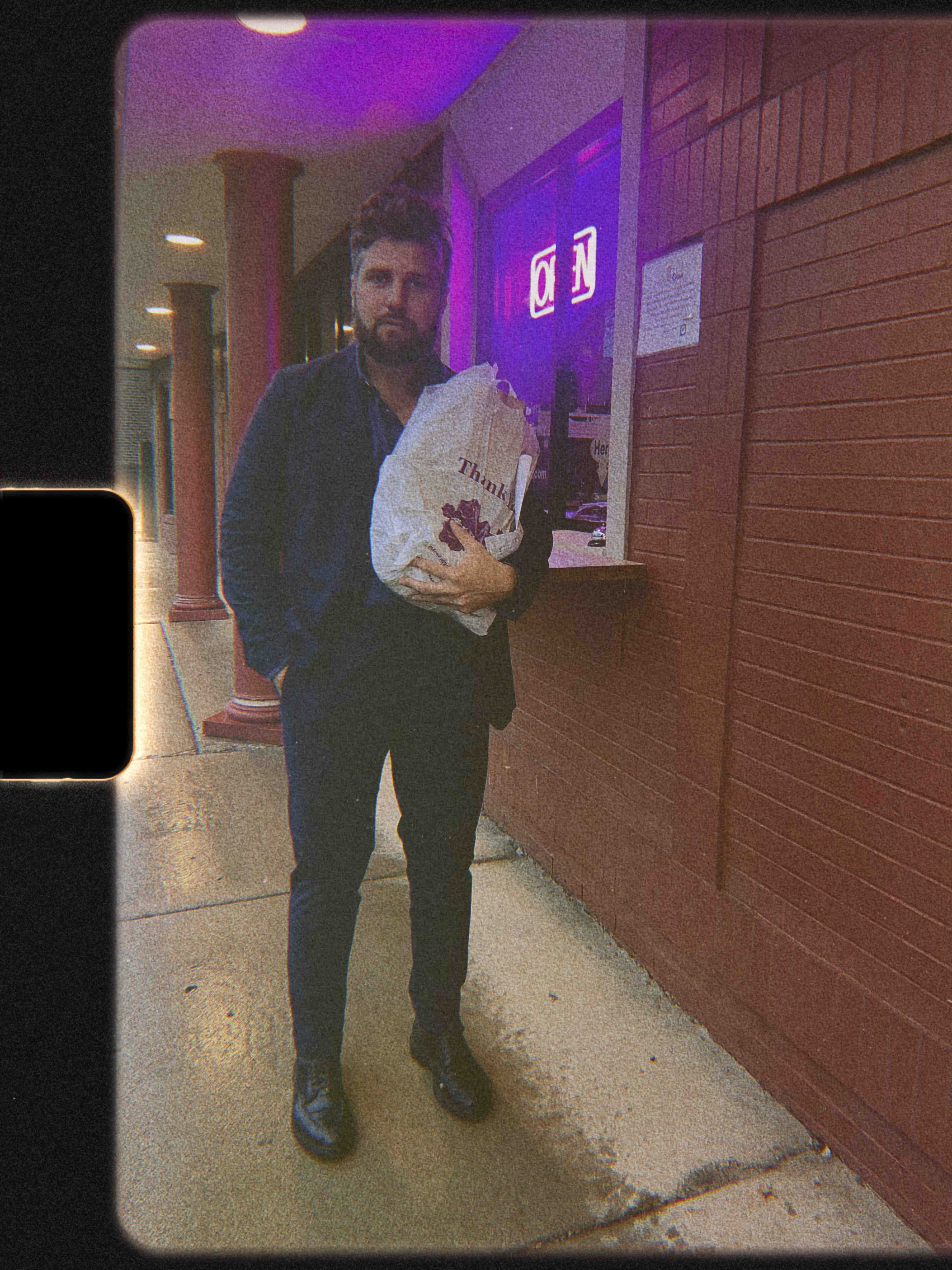 A man with dark hair and a beard stands outside at night holding a paper bag from a restaurant. He is dressed in a dark jacket, shirt, and pants. Behind him is a red wooden wall, with a sign that reads 'OPEN' in illuminated neon lights. The man is lo