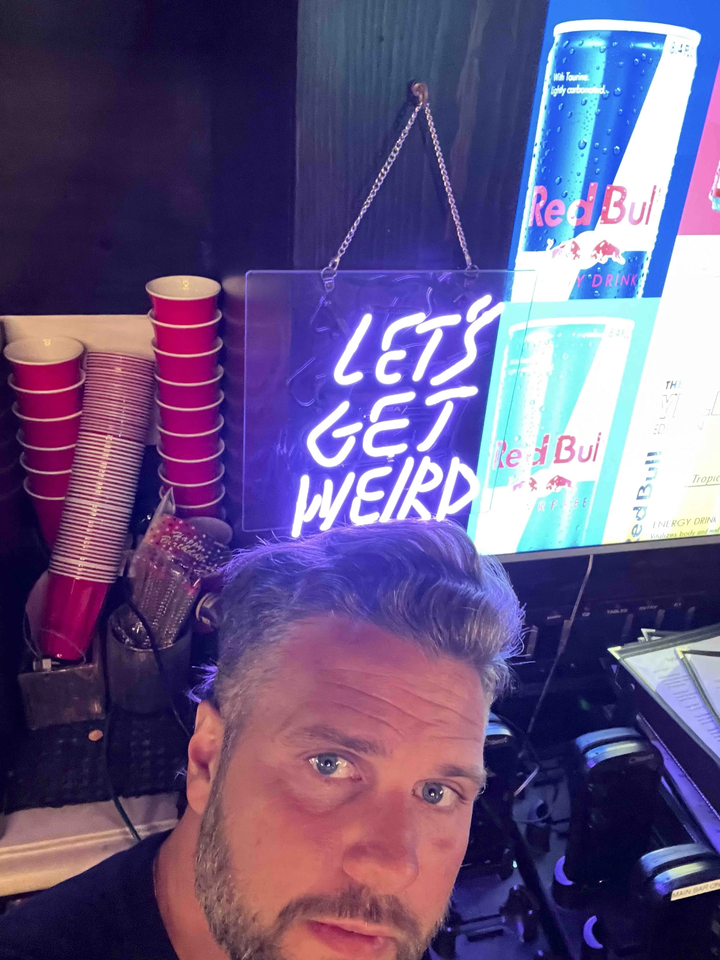 A man with gray hair and a beard takes a selfie in front of a store display. The display features a bright neon sign that says 'Let's Get Weird' and a large promotional poster for Red Bull energy drink. Several stacks of red and white cups are visibl