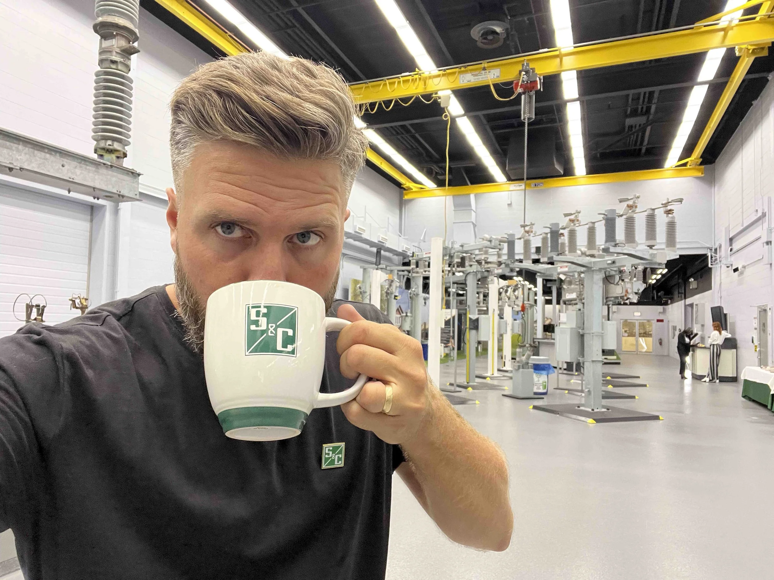 A man with light brown hair and a beard, wearing a black shirt with a green and white logo, is drinking from a white mug with a similar logo in an industrial setting with yellow overhead cranes and machinery.