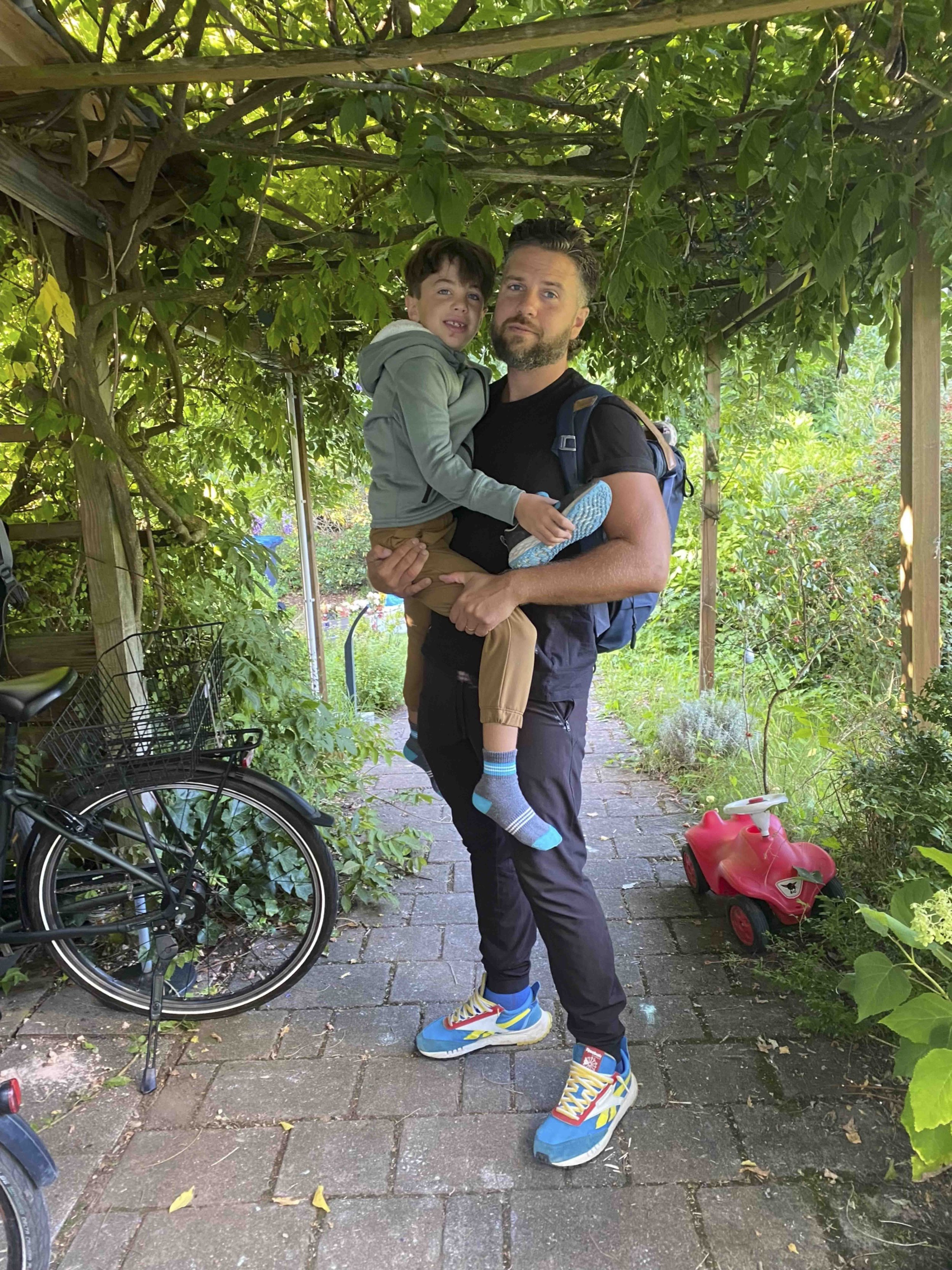 A man is holding a young boy in a garden under a wooden pergola covered with green leaves. The boy is smiling and wearing beige pants, colorful sneakers, and a light jacket, while the man is dressed in black pants, a black shirt, and colorful sneaker