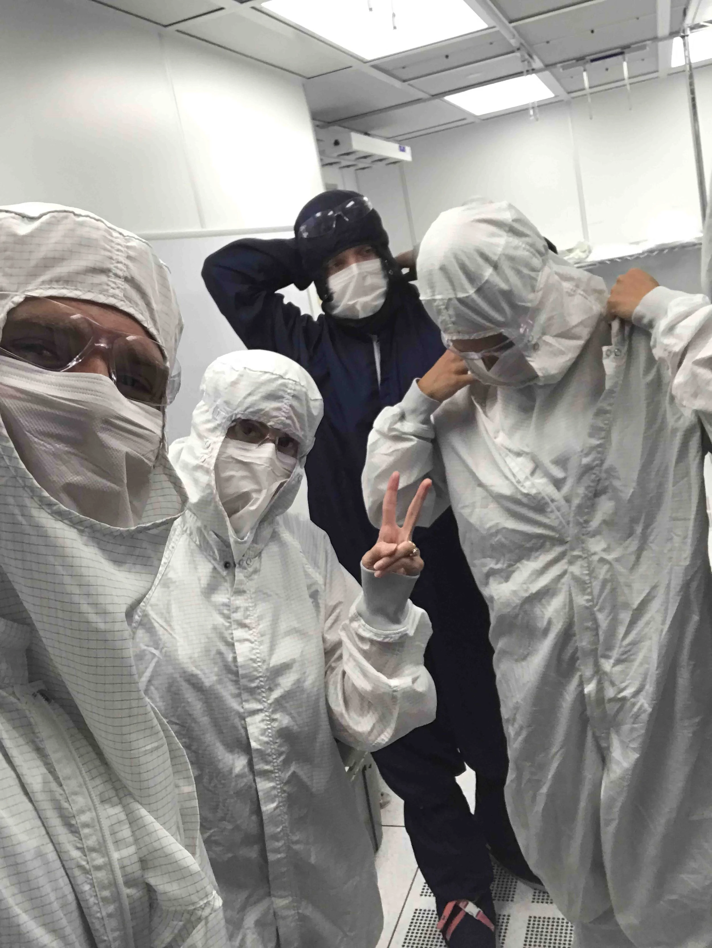 People wearing white protective suits, masks, and goggles inside an elevator, with a person in black holding their head and another person making a peace sign.