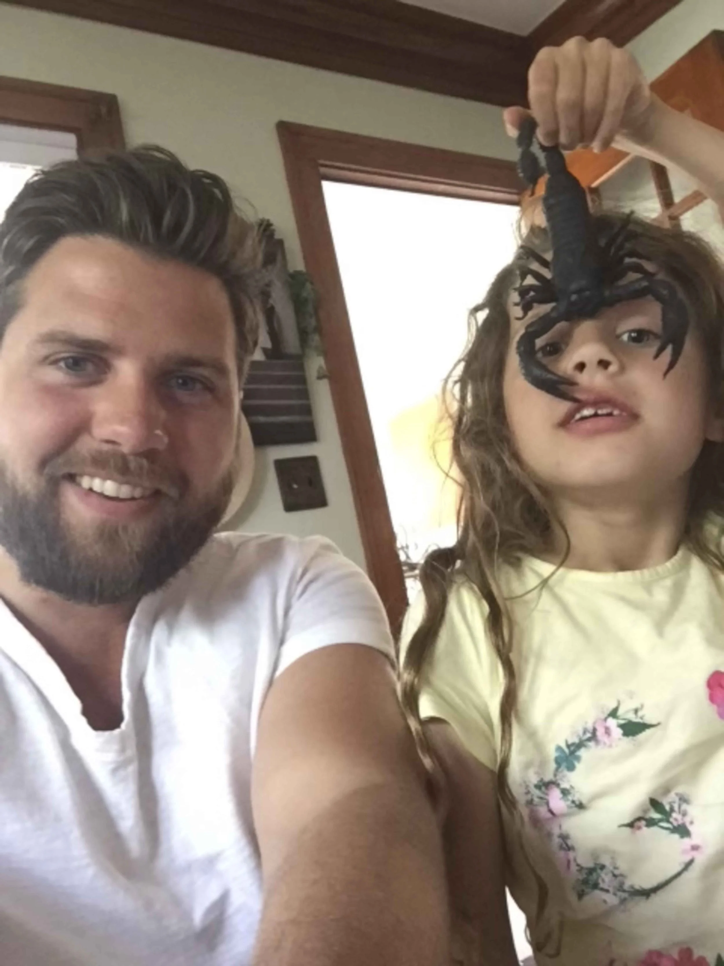 A man and a girl taking a selfie inside a house, with the girl holding a large spider on her hand.