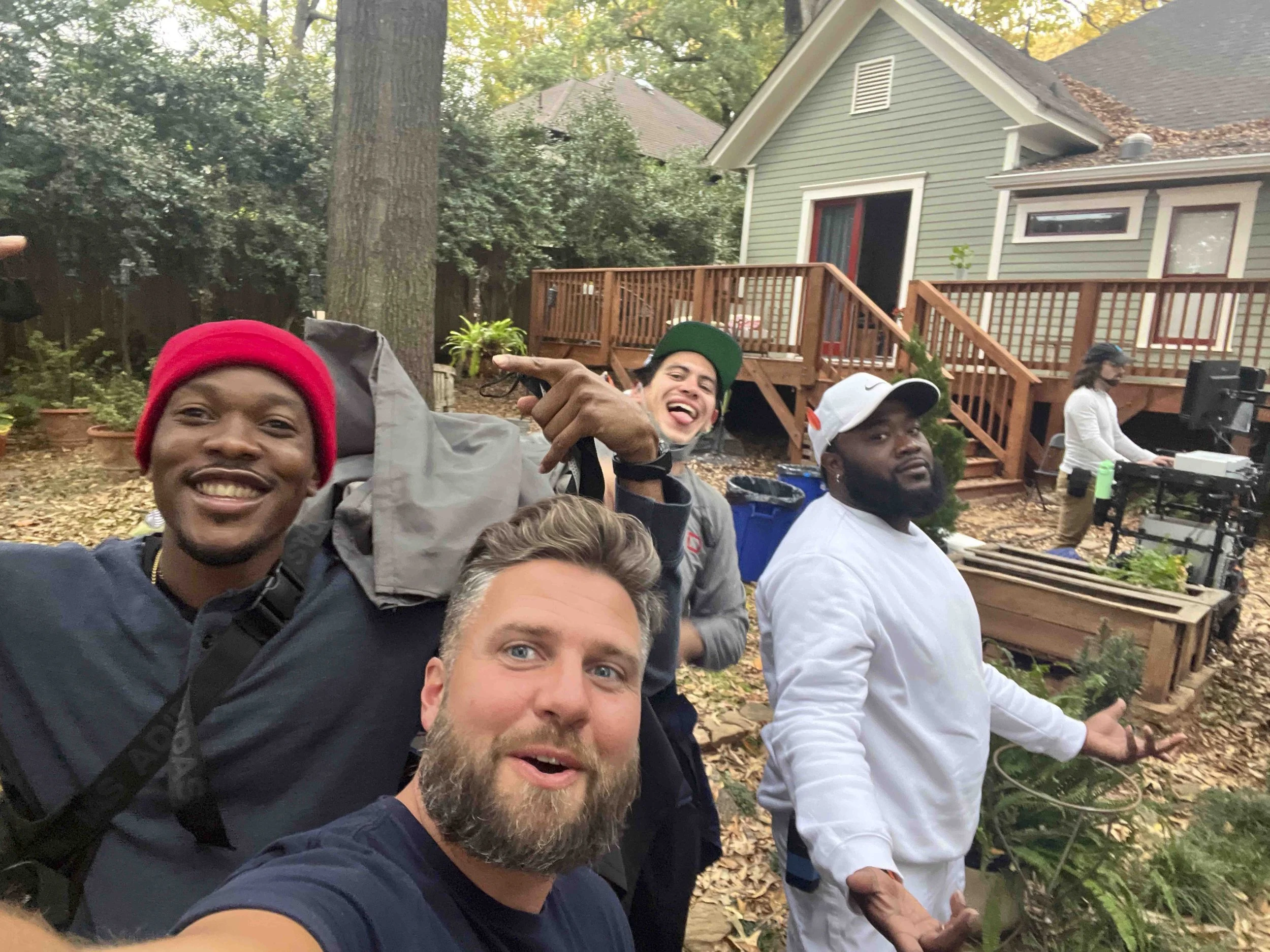 Group of people smiling and posing for a selfie outdoors in front of a house with a wooden deck and trees, some wearing hats and face masks.