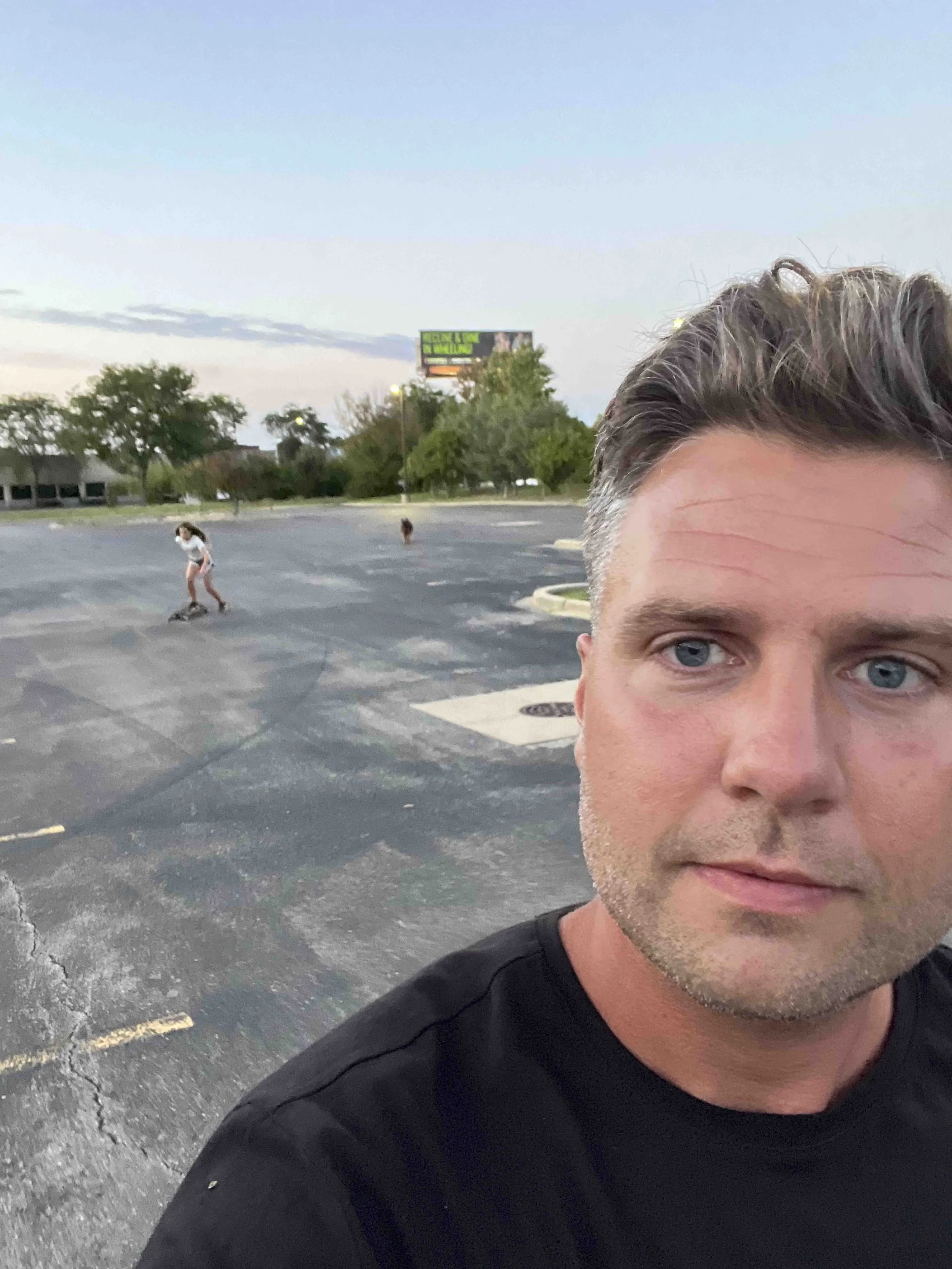 Close-up of a man's face with blue eyes, face partially visible, black shirt, in an empty parking lot with a girl skateboarding in the background, trees, and a billboard in the distance.