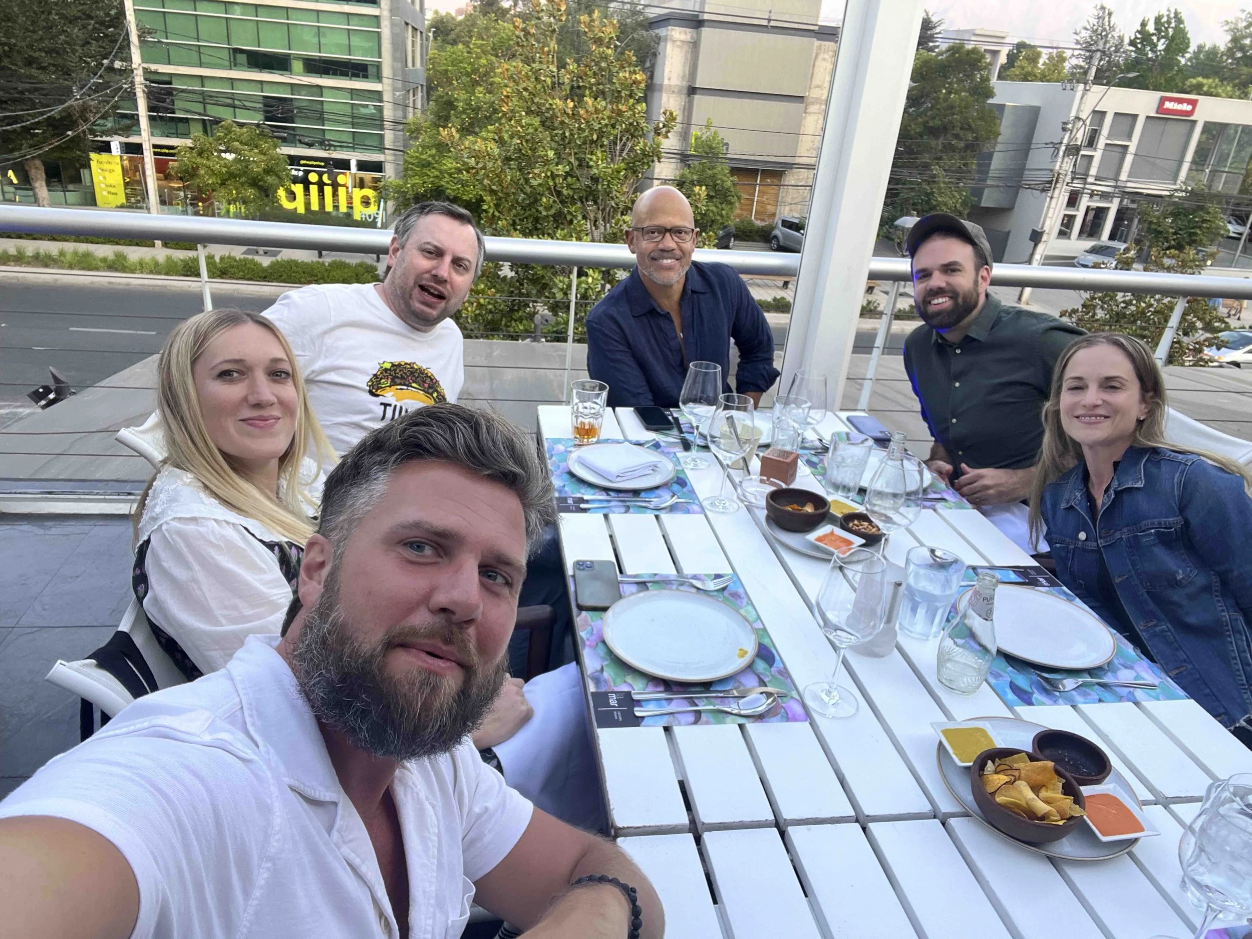 Six people sitting around a dining table outdoors on a patio, with city buildings and trees in the background, enjoying a meal together.