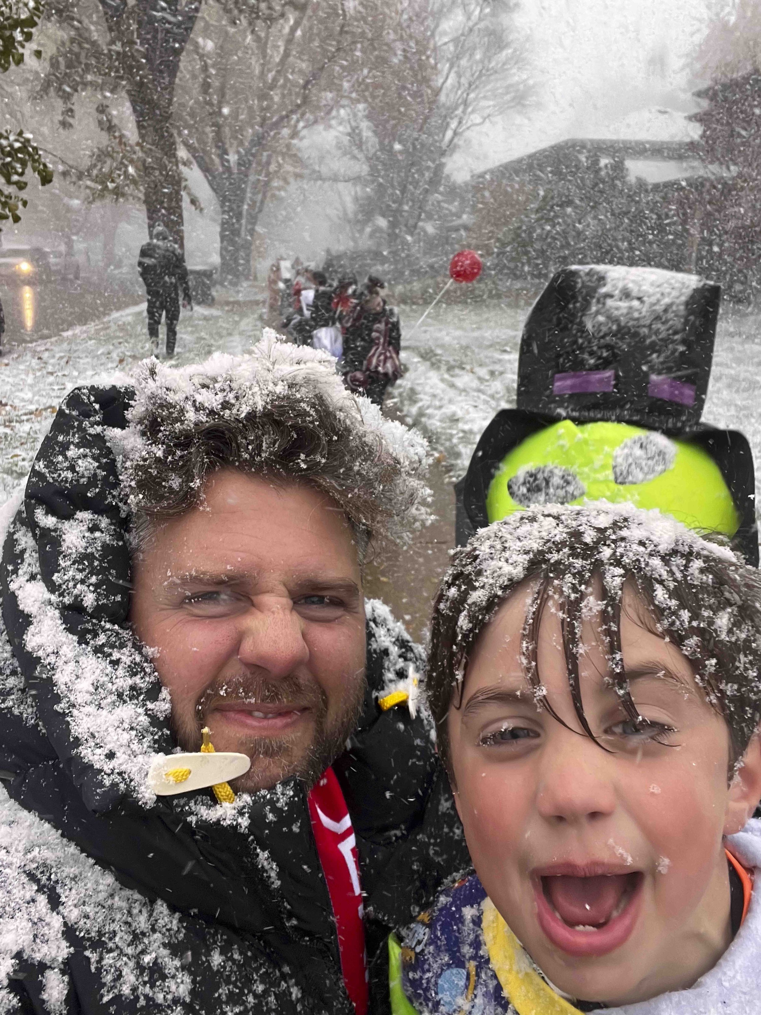 A man and a young boy smiling and enjoying a snowstorm, with snow covering their faces and hair, during a snowy day outdoors.