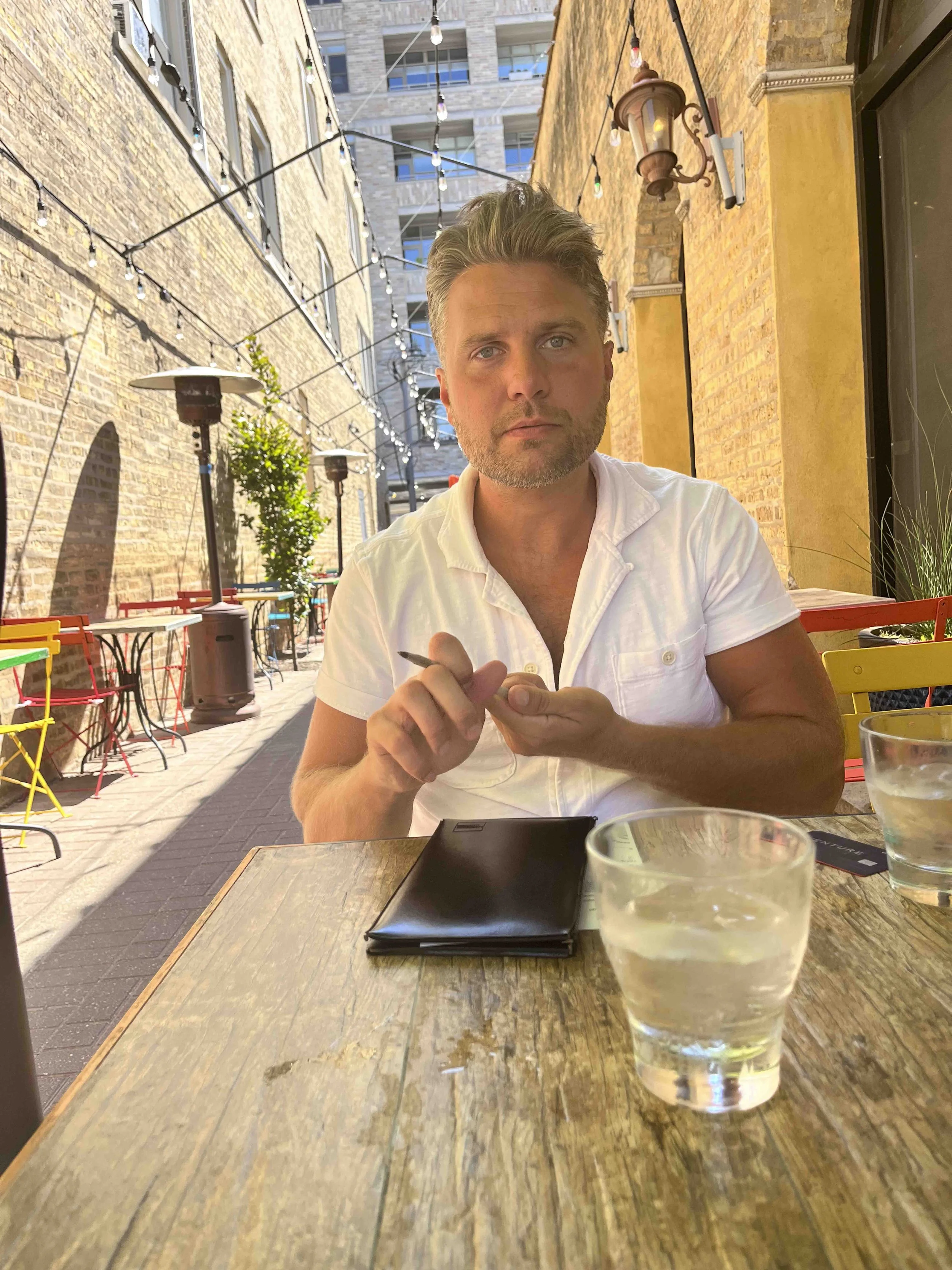 A man with light brown hair and a beard sitting at a wooden table outdoors in a city setting with brick walls and string lights overhead. He is wearing a white shirt and holding a pen, with a black tablet and a glass of water on the table.