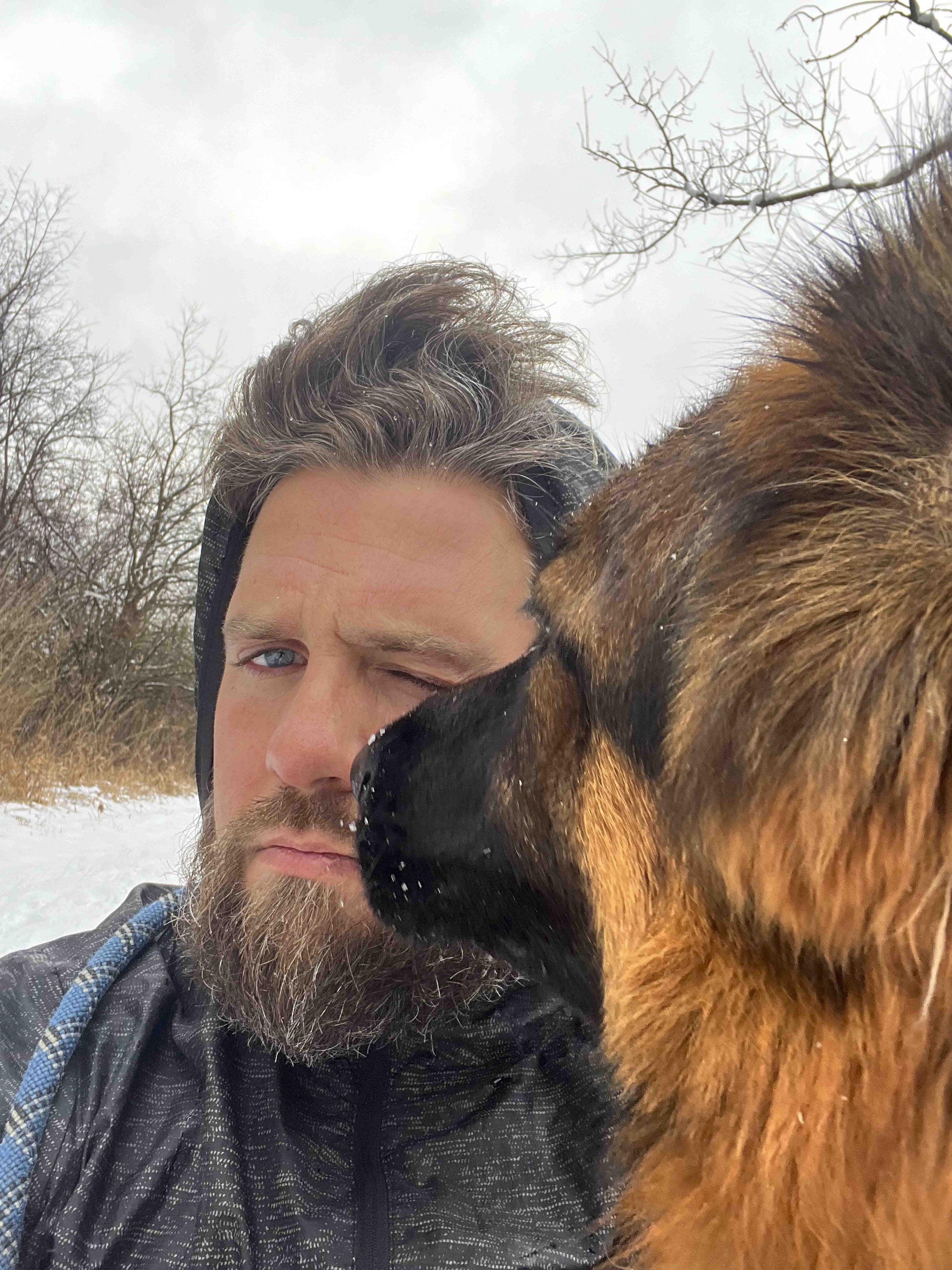 A person with gray hair, partly visible face and blue eyes, taking a selfie with a large brown dog outdoors in a snowy, leafless wooded area.