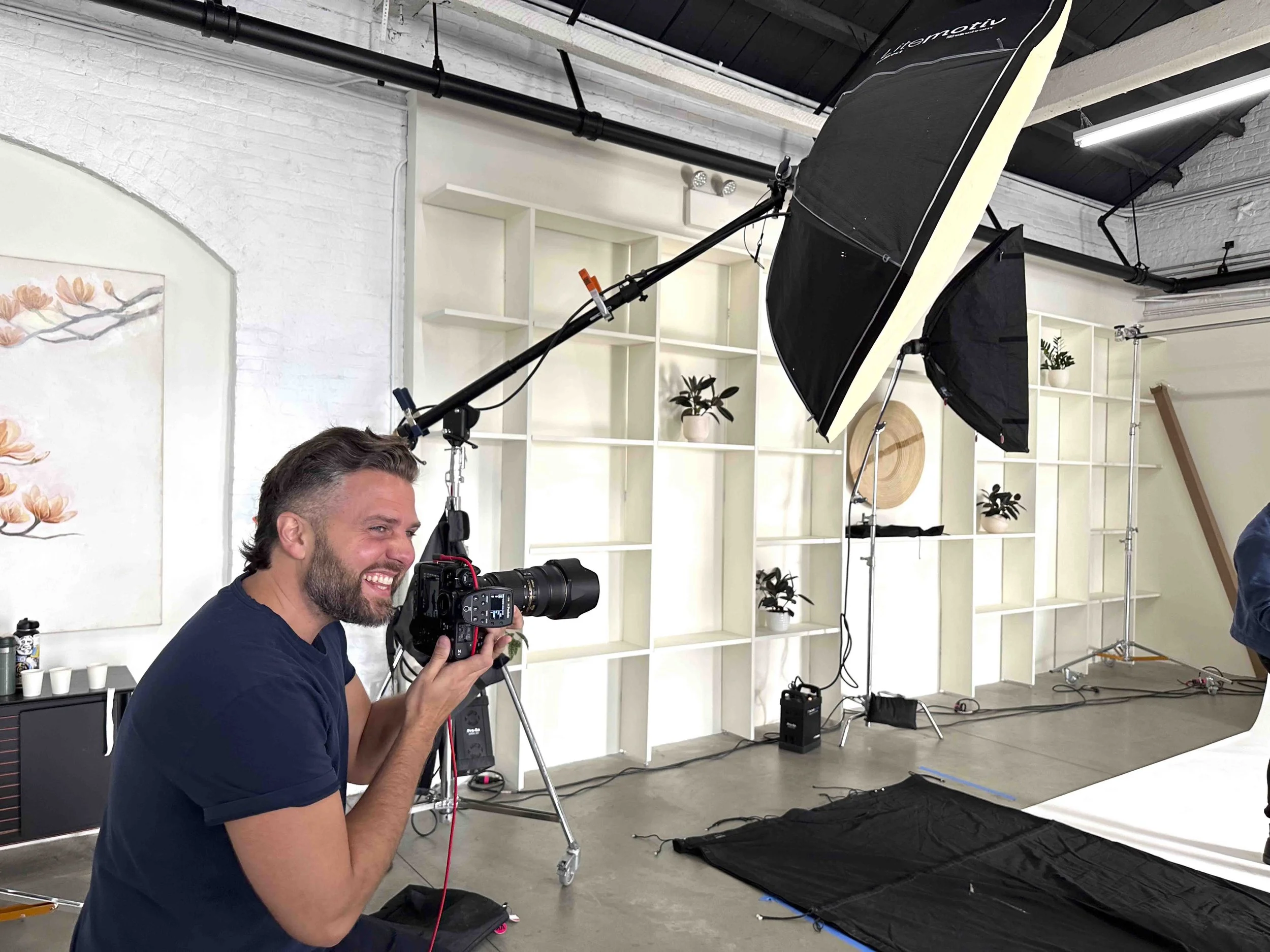 A man with dark hair and a beard is smiling and holding a professional camera with a large lens in a photography studio. The studio has white brick walls, shelves with potted plants, and professional lighting equipment including a softbox and umbrell