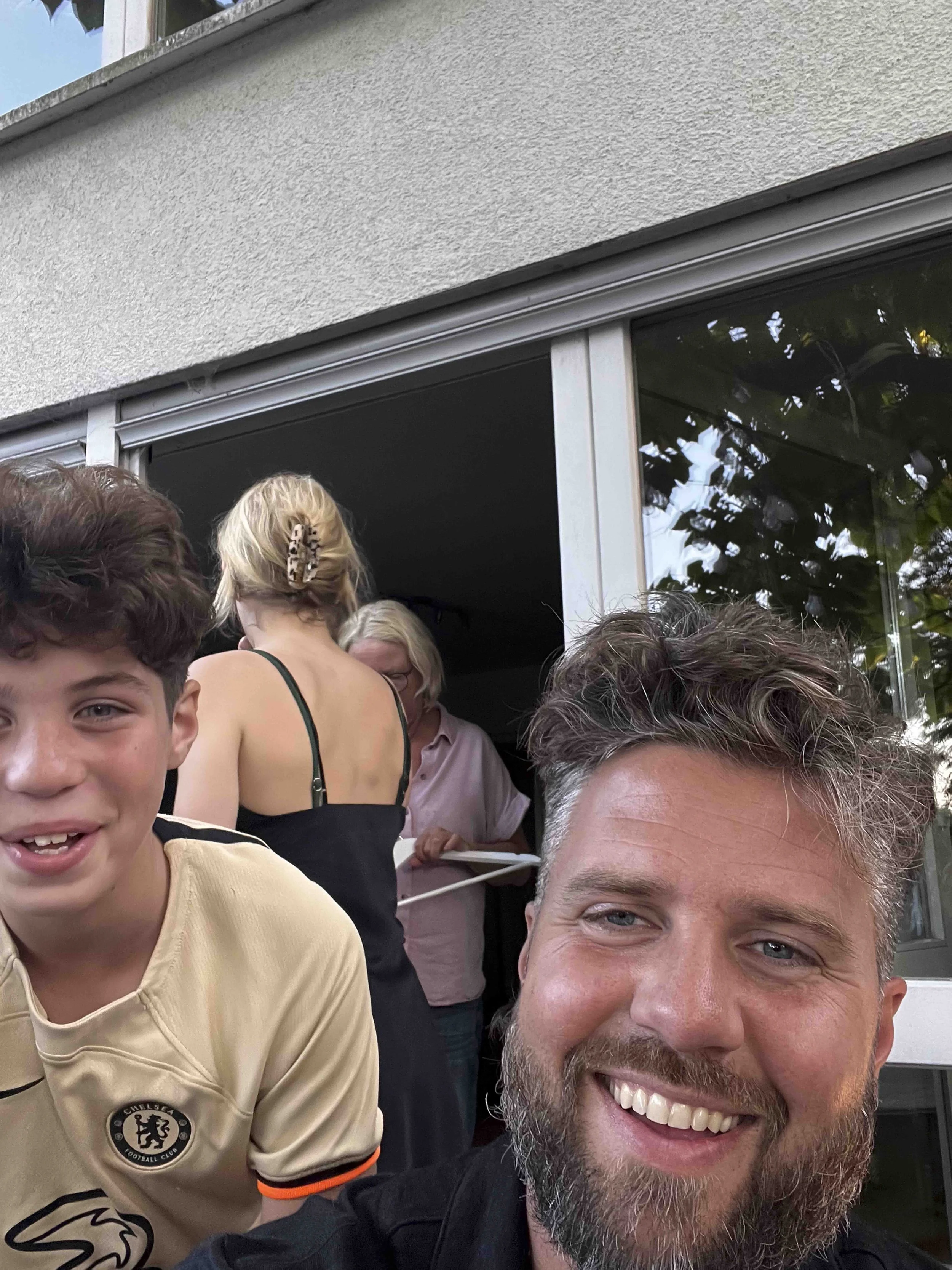 Smiling man with a beard taking a selfie with a boy wearing a Chelsea Football Club jersey, surrounded by people gathered outdoors near a building with a large window.